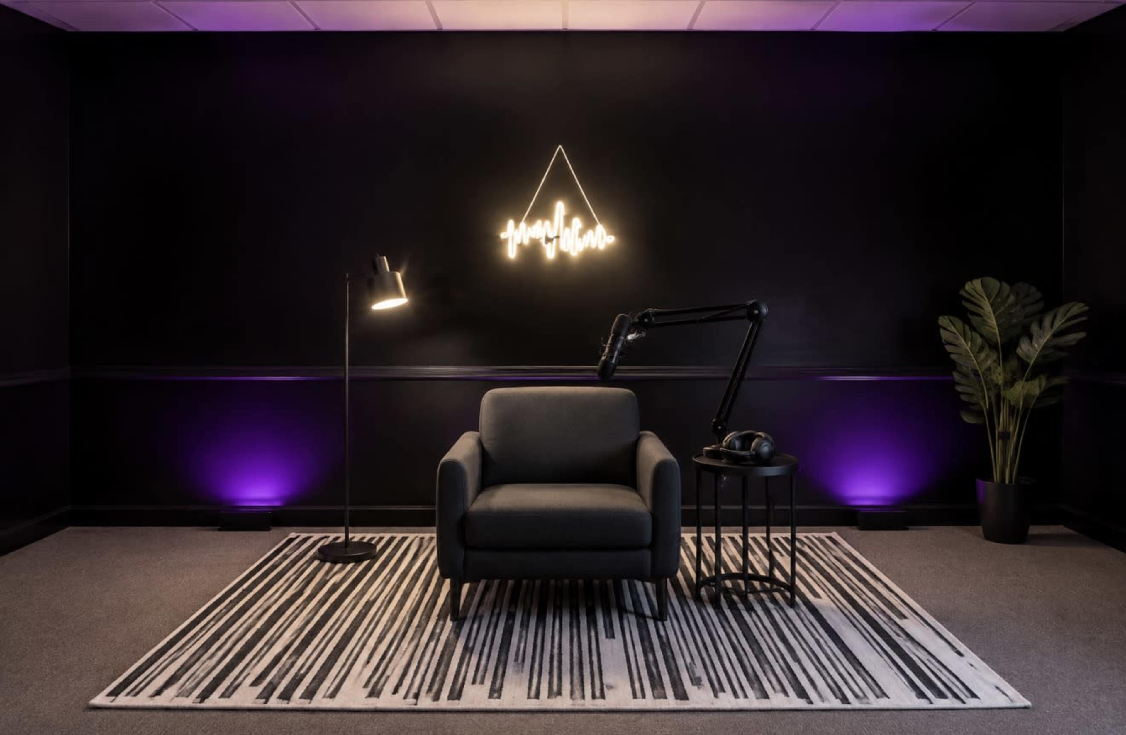 The image shows a modern lounge area with a black wall, a single gray armchair on a striped rug, a floor lamp, a small table, and a large potted plant, all illuminated by purple lighting.