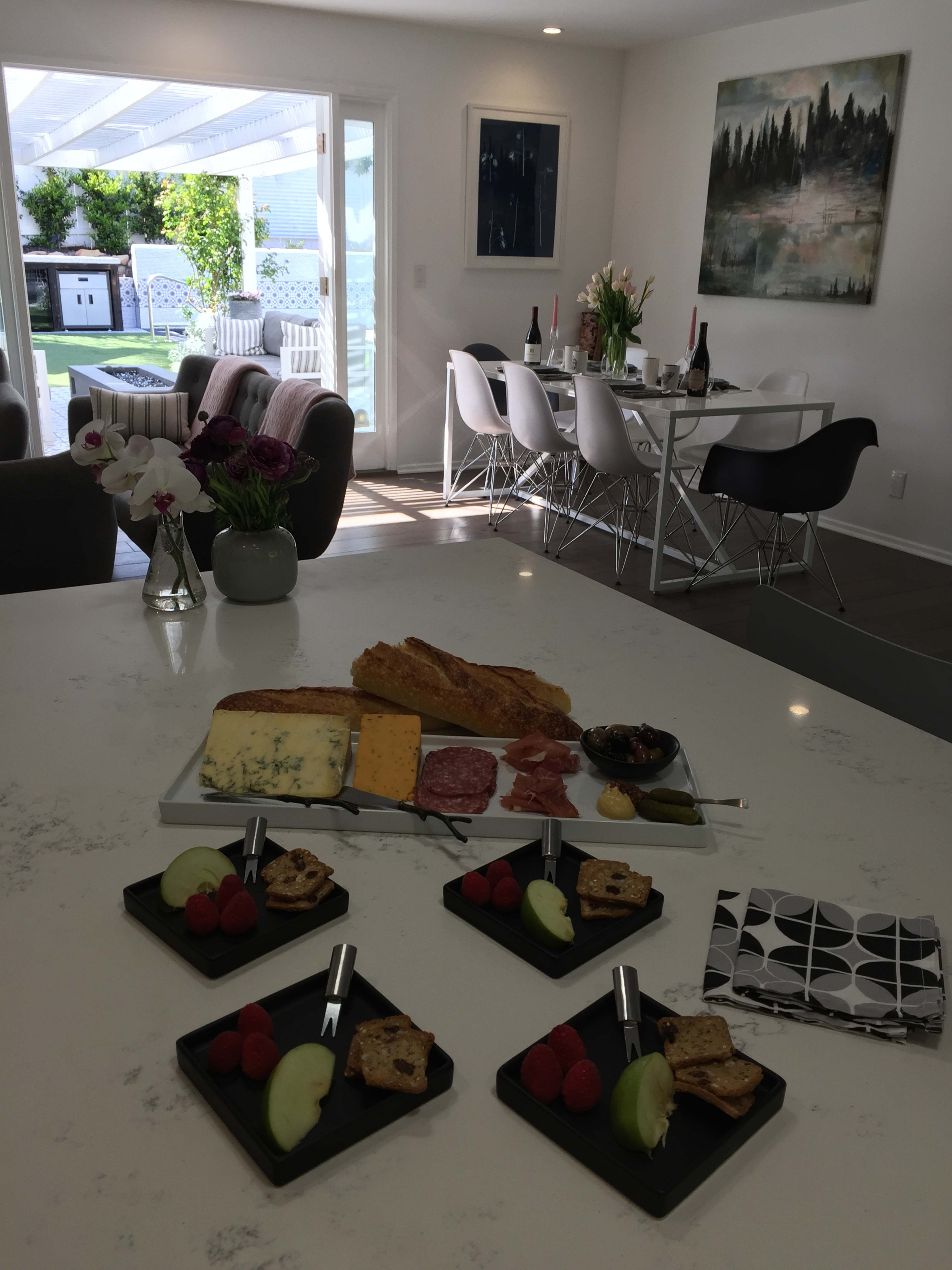 A kitchen counter is set with a charcuterie board, featuring assorted cheeses, meats, and fruits, while a dining area is visible in the background with a table set for a meal.