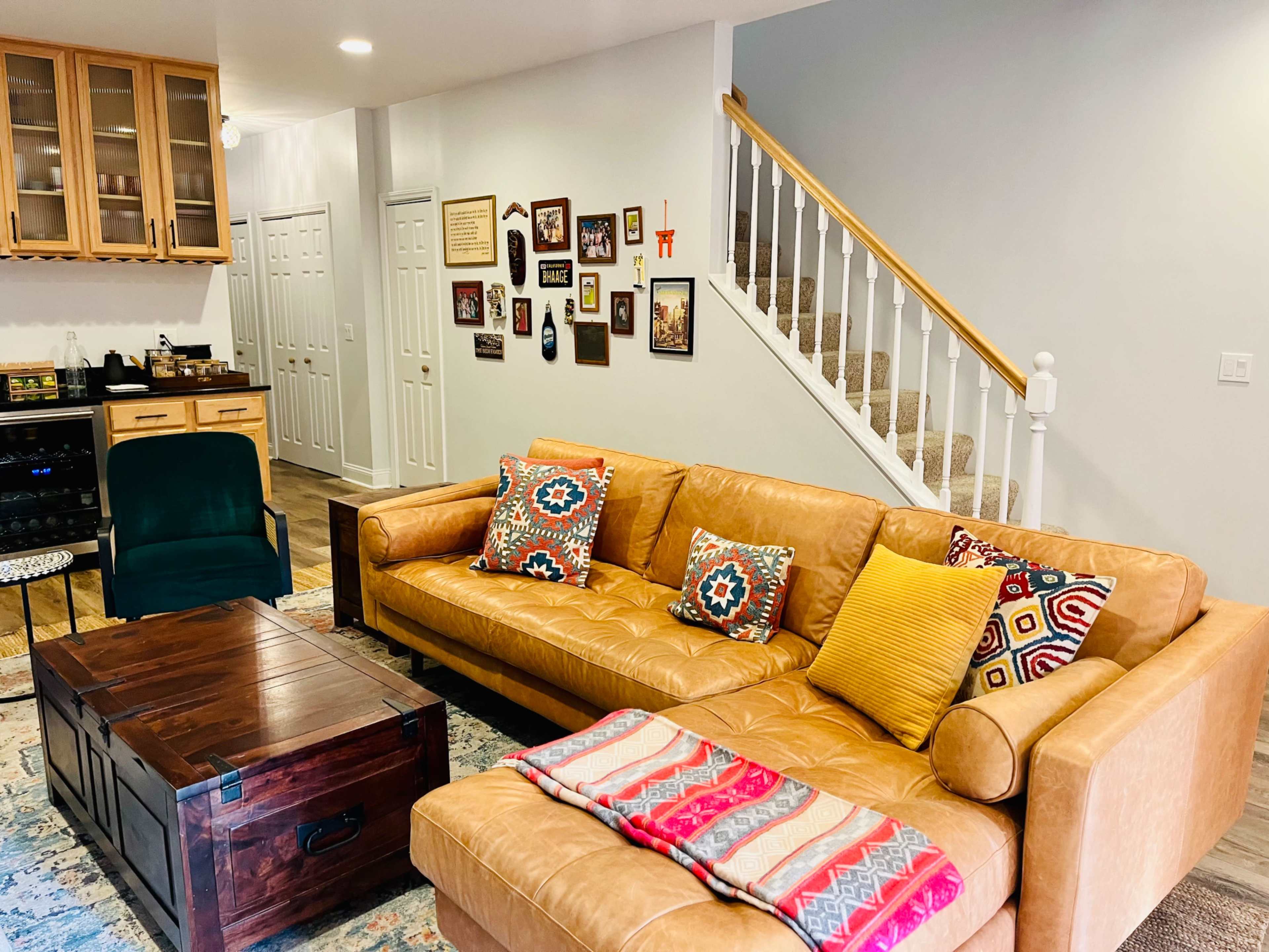 A cozy living room features a leather sofa with patterned pillows, a wooden coffee table, and a collection of framed pictures on the wall, with a staircase visible in the background.