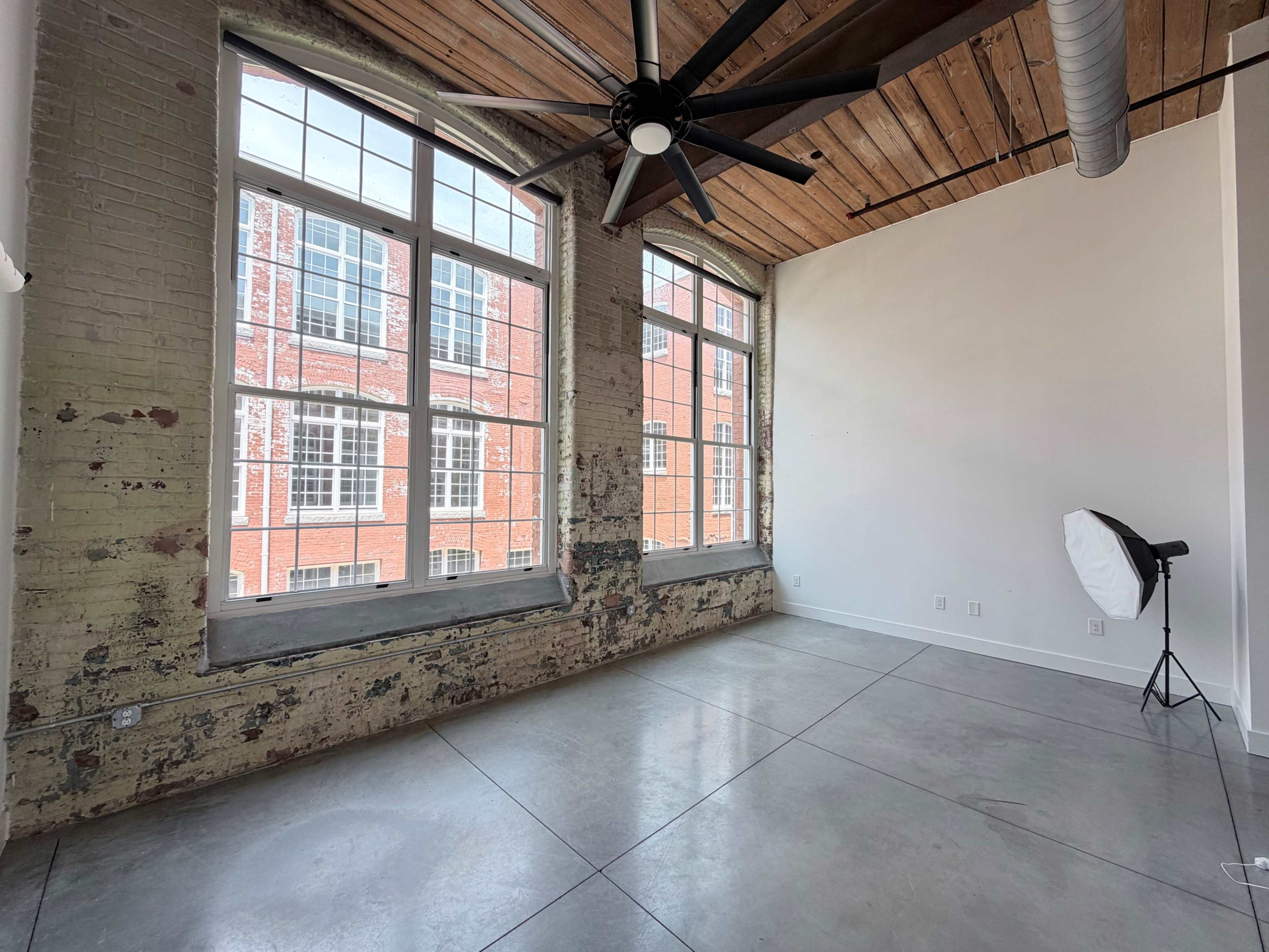 The image shows an expansive room with large windows, exposed brick walls, and concrete floors, featuring a ceiling fan and a lighting setup in one corner.