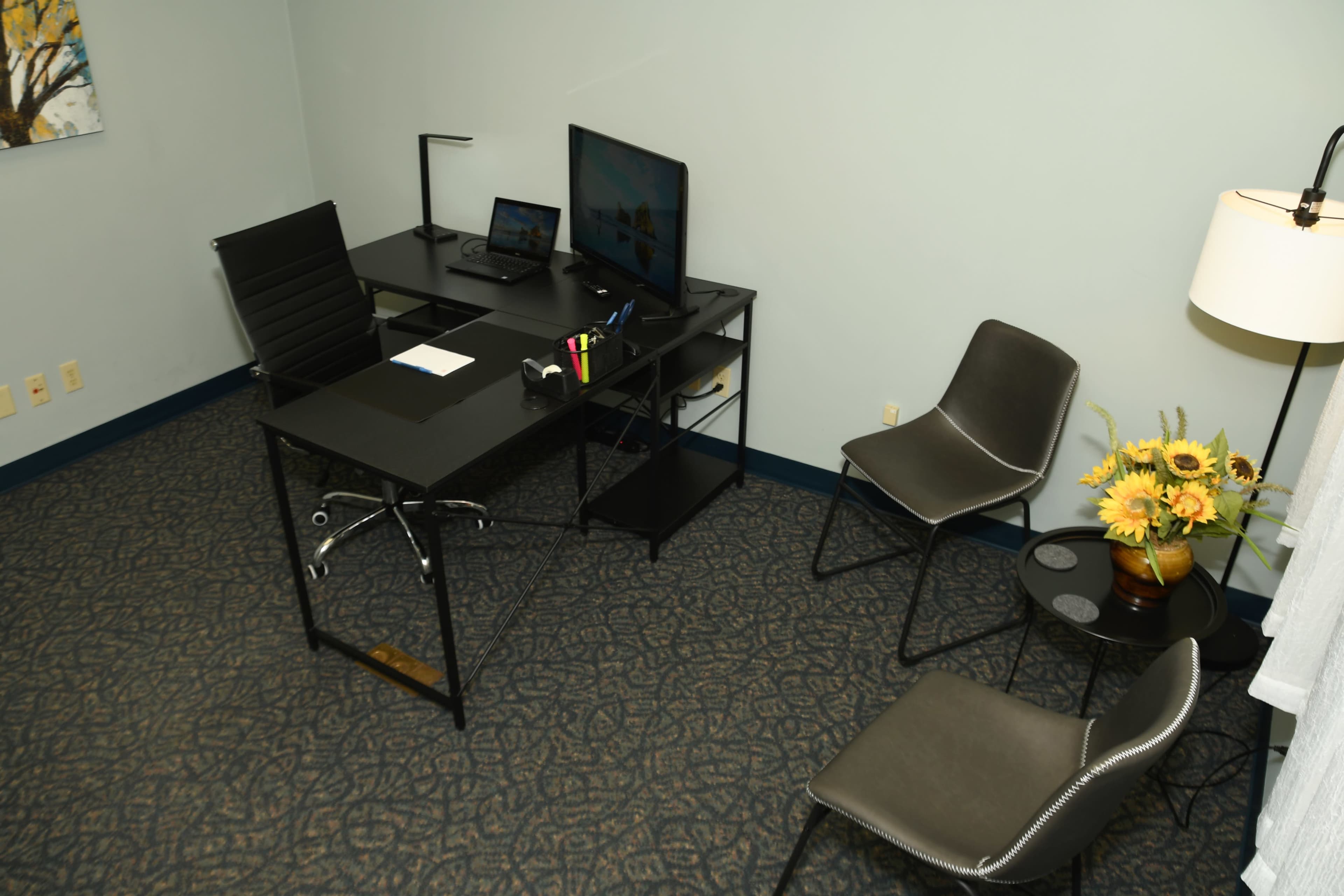 The image shows a minimalistic office space featuring a black desk with a computer monitor, a chair, and two additional chairs arranged nearby, along with a small table holding a vase of flowers.