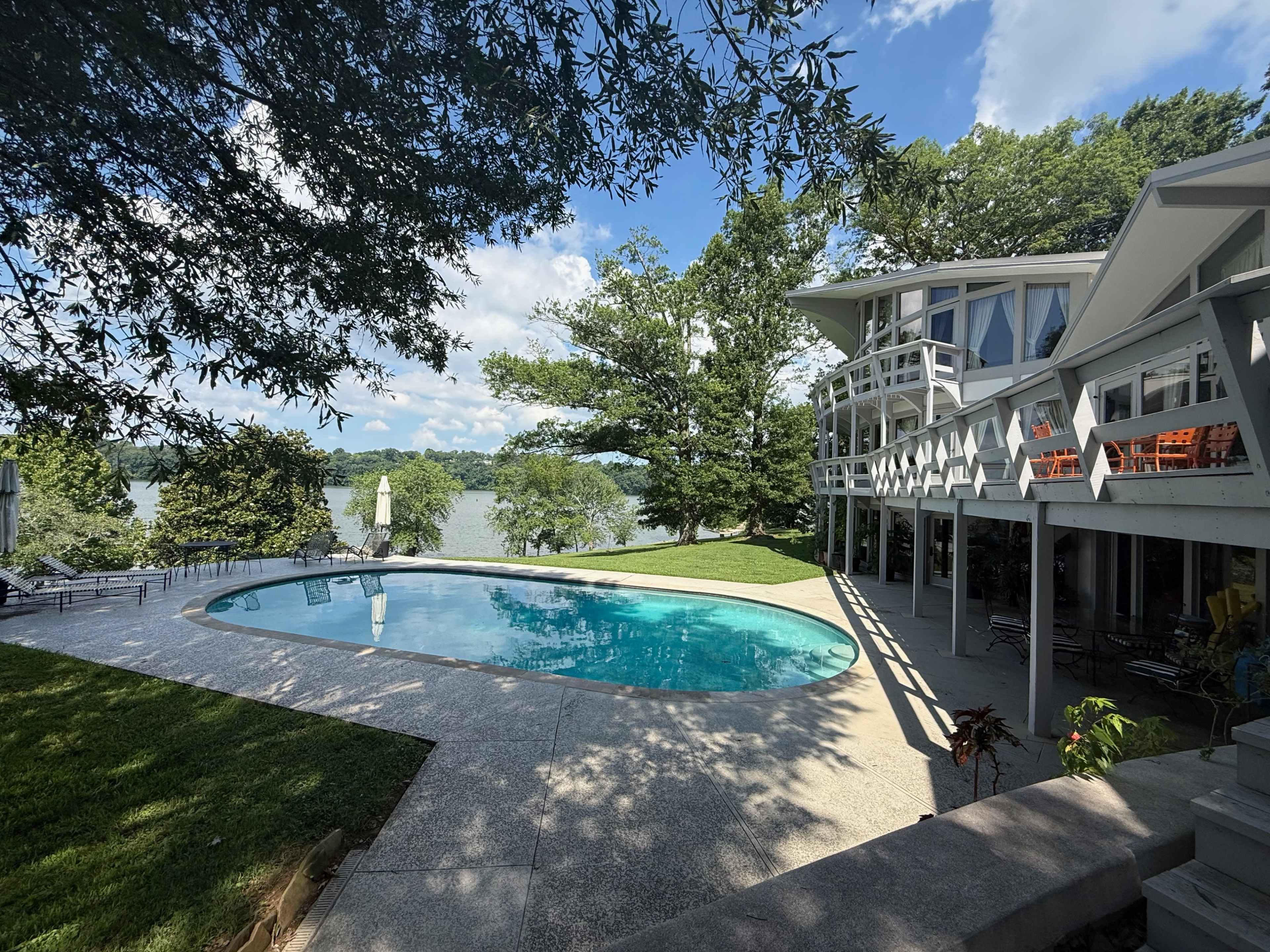 The image shows a modern house with a large swimming pool and a lush green lawn next to a lake, surrounded by trees under a partly cloudy sky.