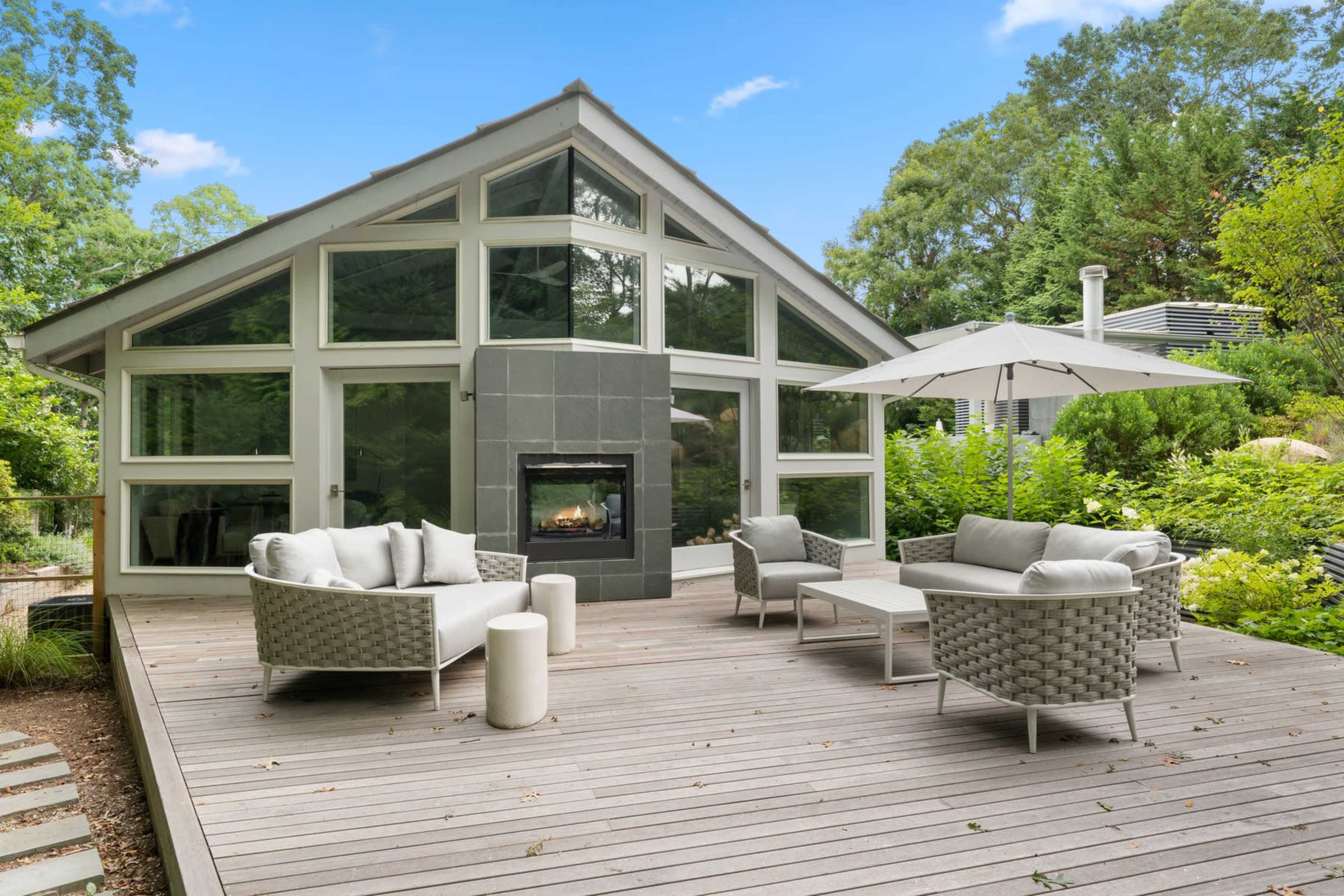 A modern deck features comfortable seating arrangements and a patio umbrella in front of a large glass-paneled house surrounded by lush greenery.