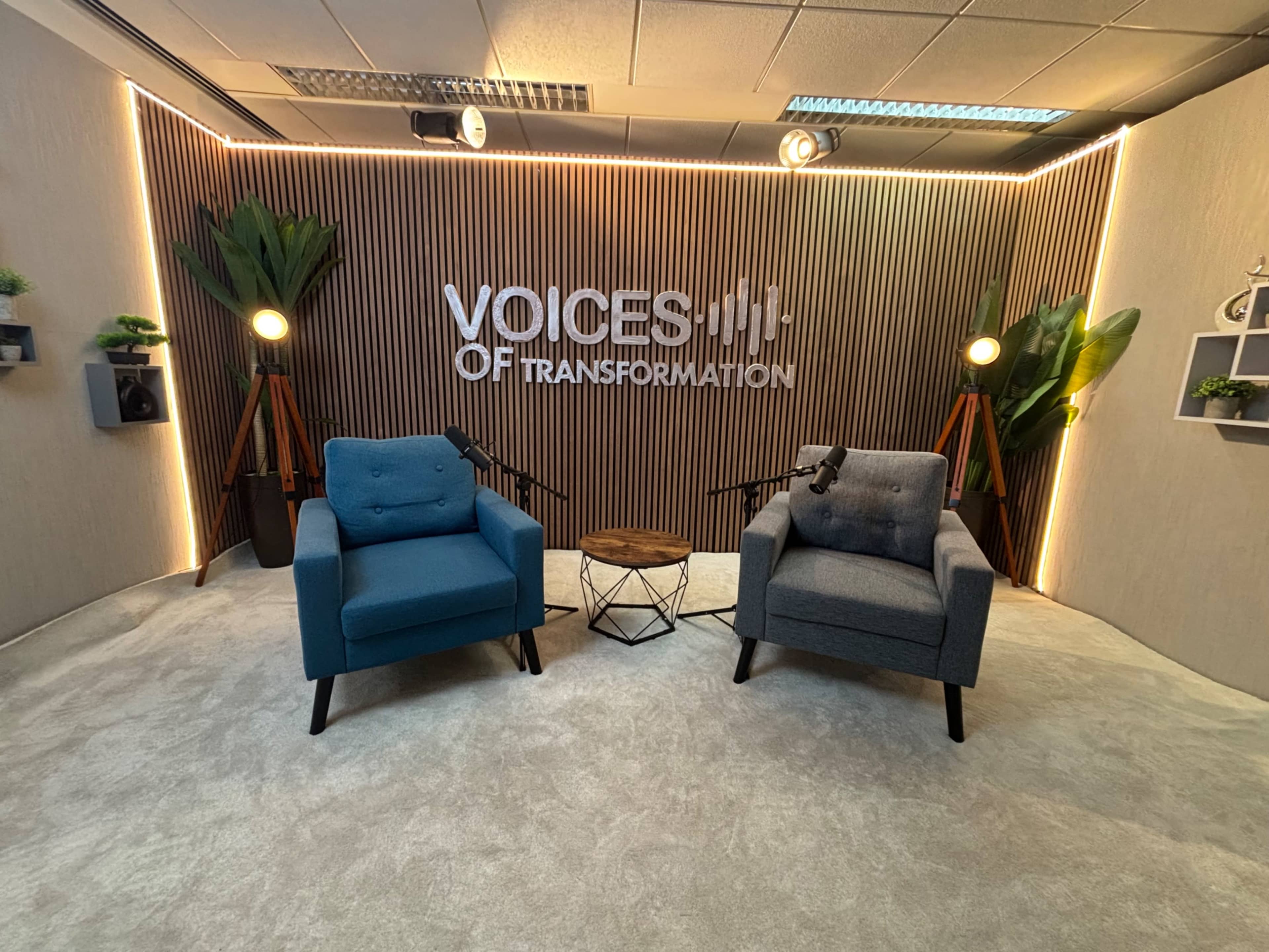 The image shows a modern studio setup with two armchairs, a small table, and a backdrop featuring the text "VOICES OF TRANSFORMATION," illuminated with soft lighting and surrounded by decorative plants.