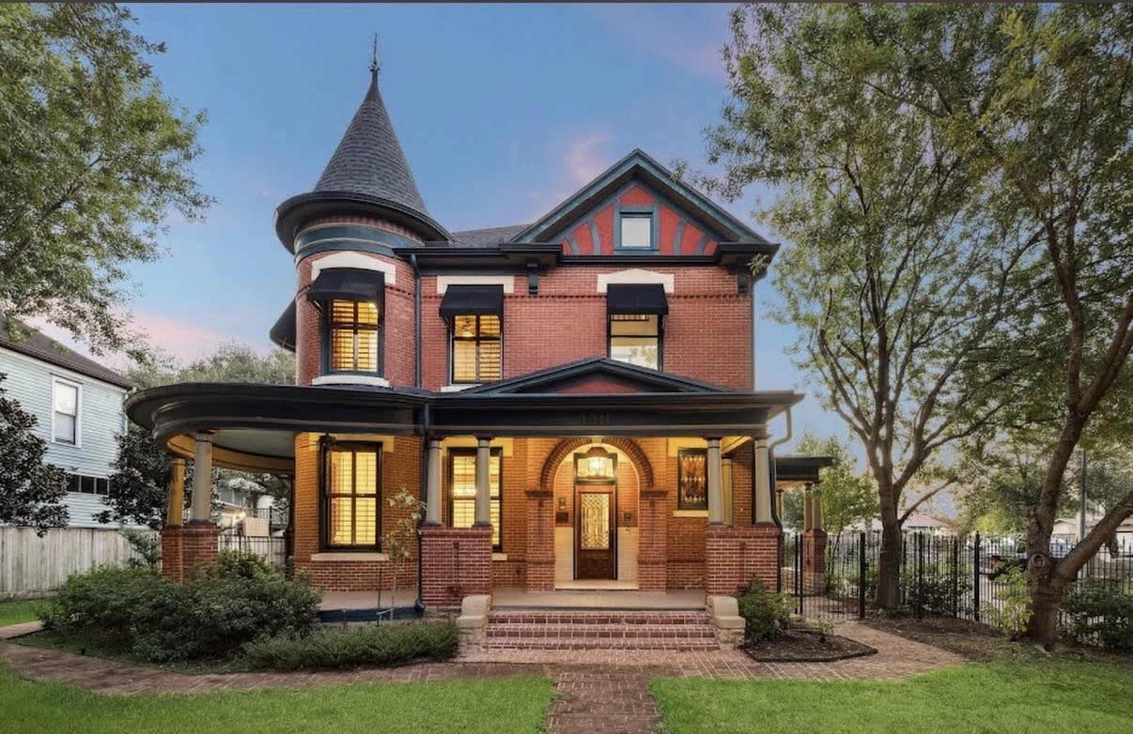 The image shows a large, Victorian-style house with a turret and a wrap-around porch, surrounded by landscaped greenery.