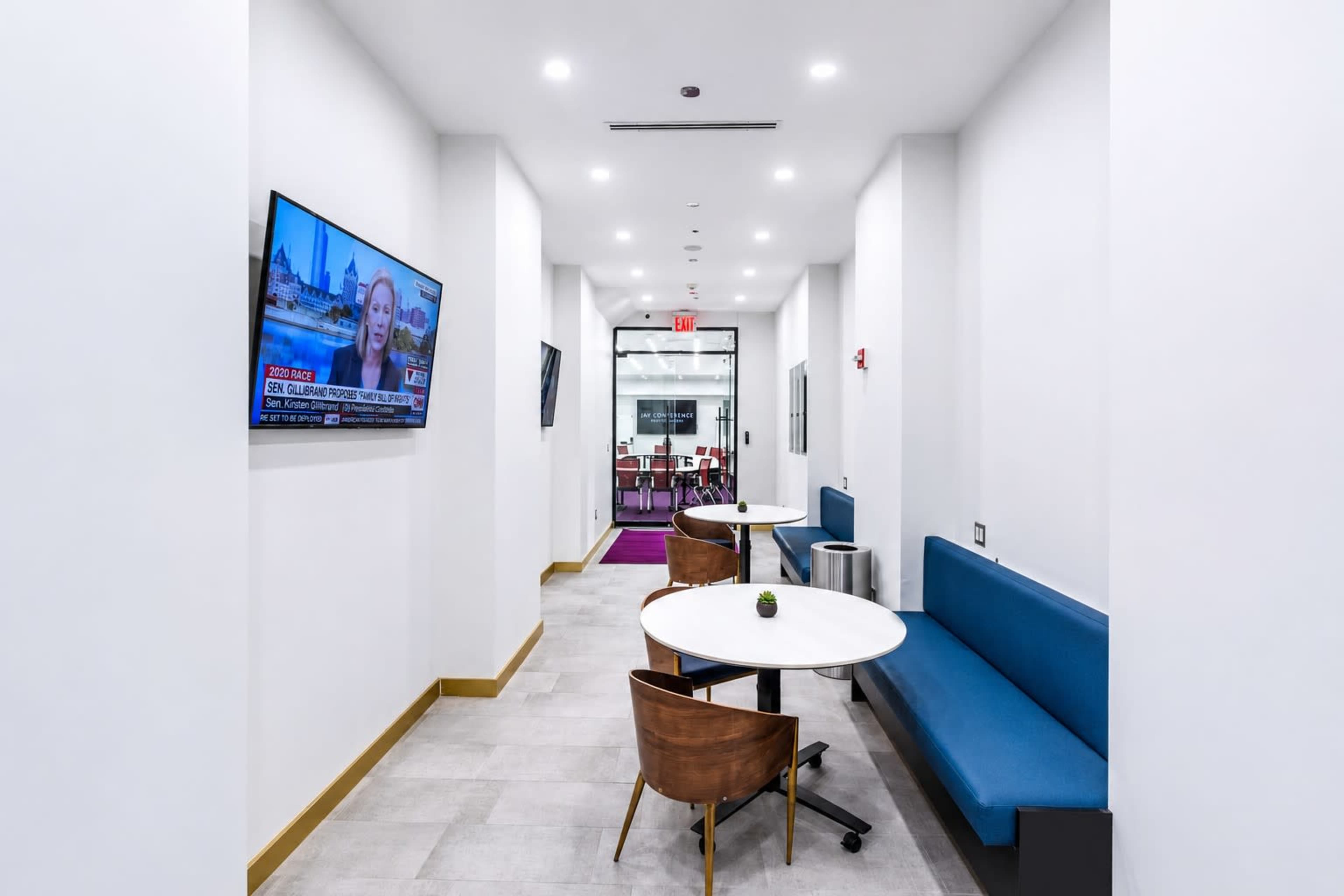 The image shows a bright, modern hallway with a television displaying news, two tables with chairs, and a glass door leading to another room.