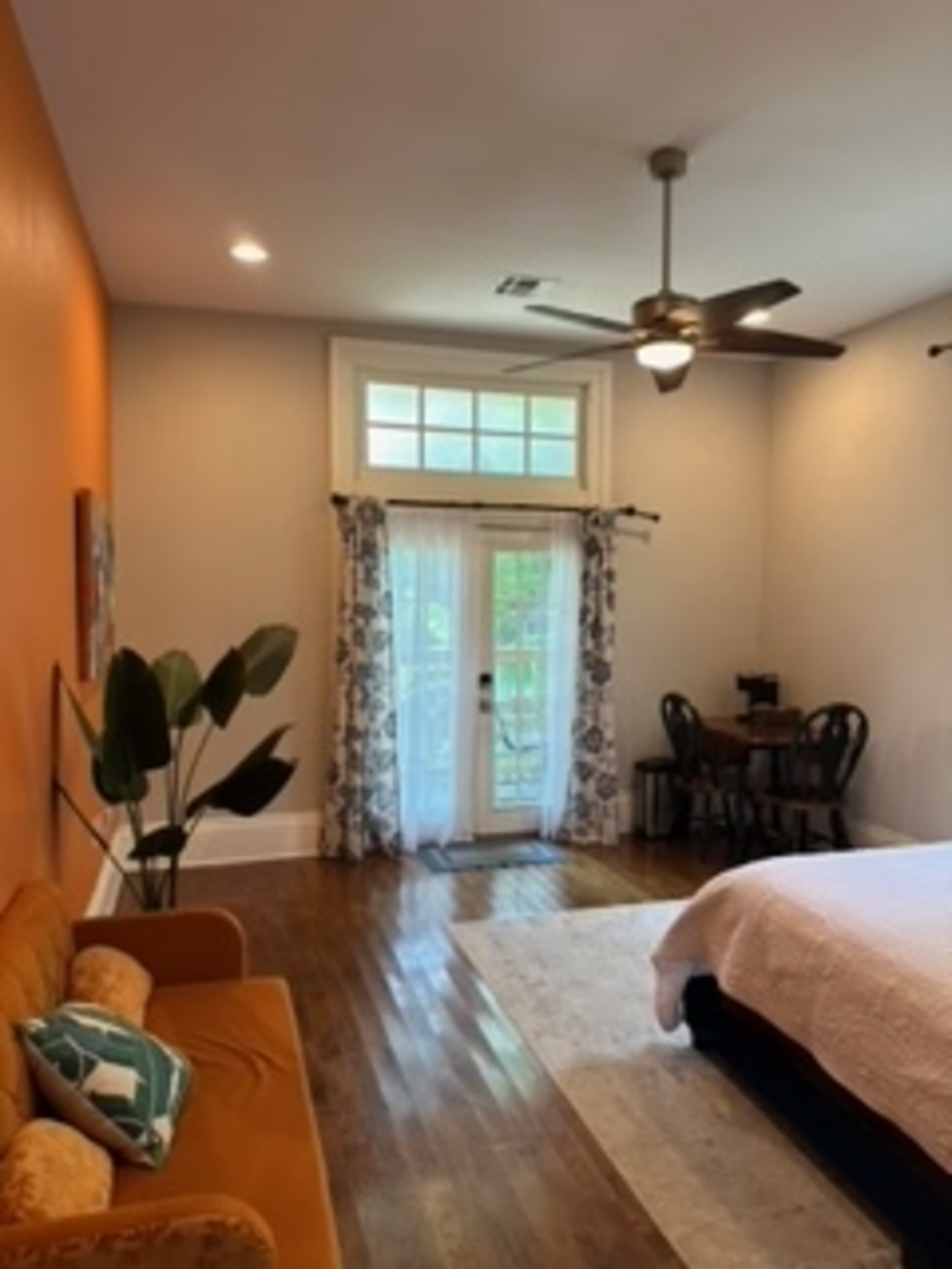 The image shows a spacious bedroom featuring a queen-sized bed, a small dining area with two chairs, and a door leading to an outdoor balcony.