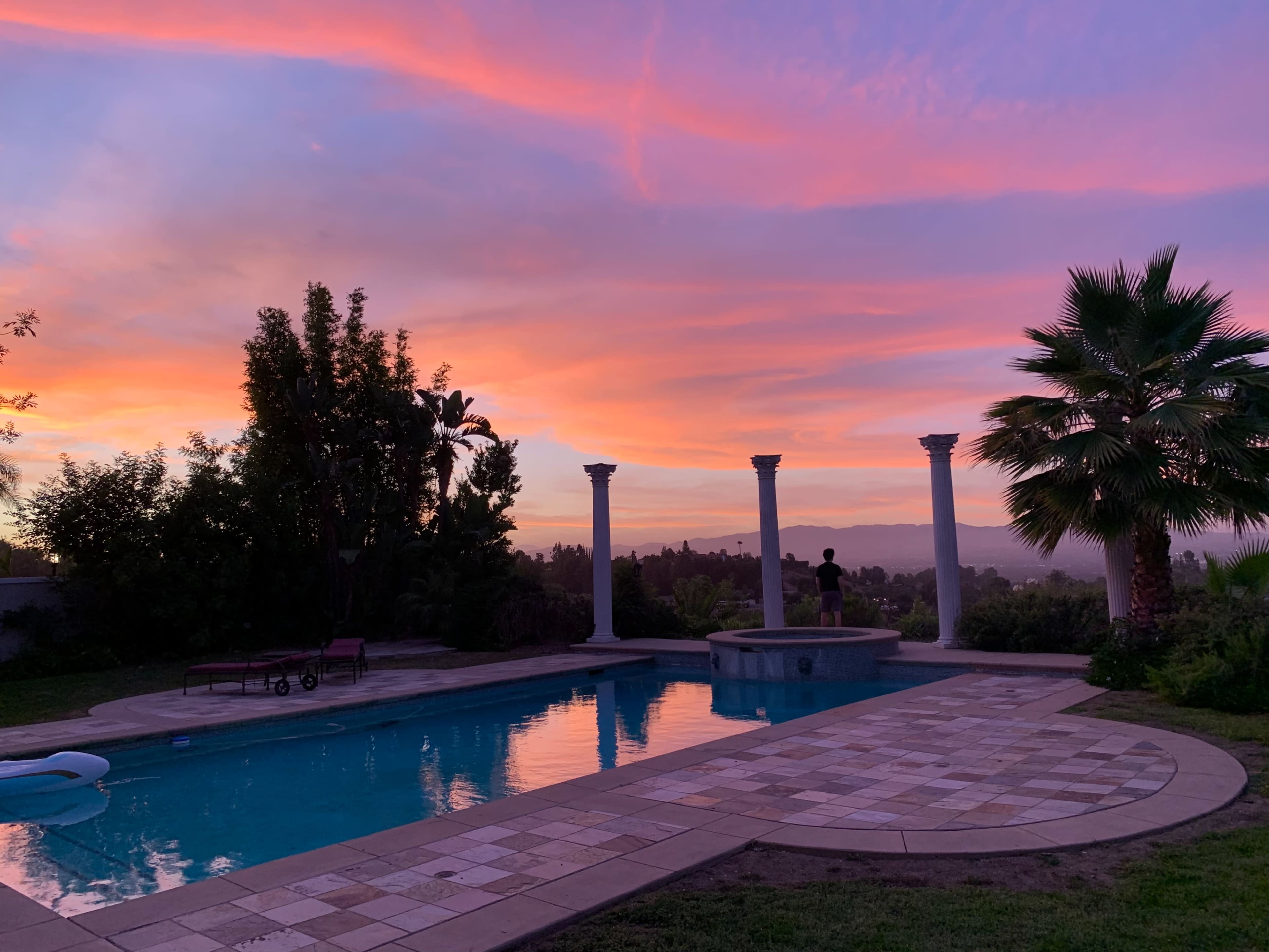 A person stands by a swimming pool, silhouetted against a vibrant sunset sky with shades of pink and orange.