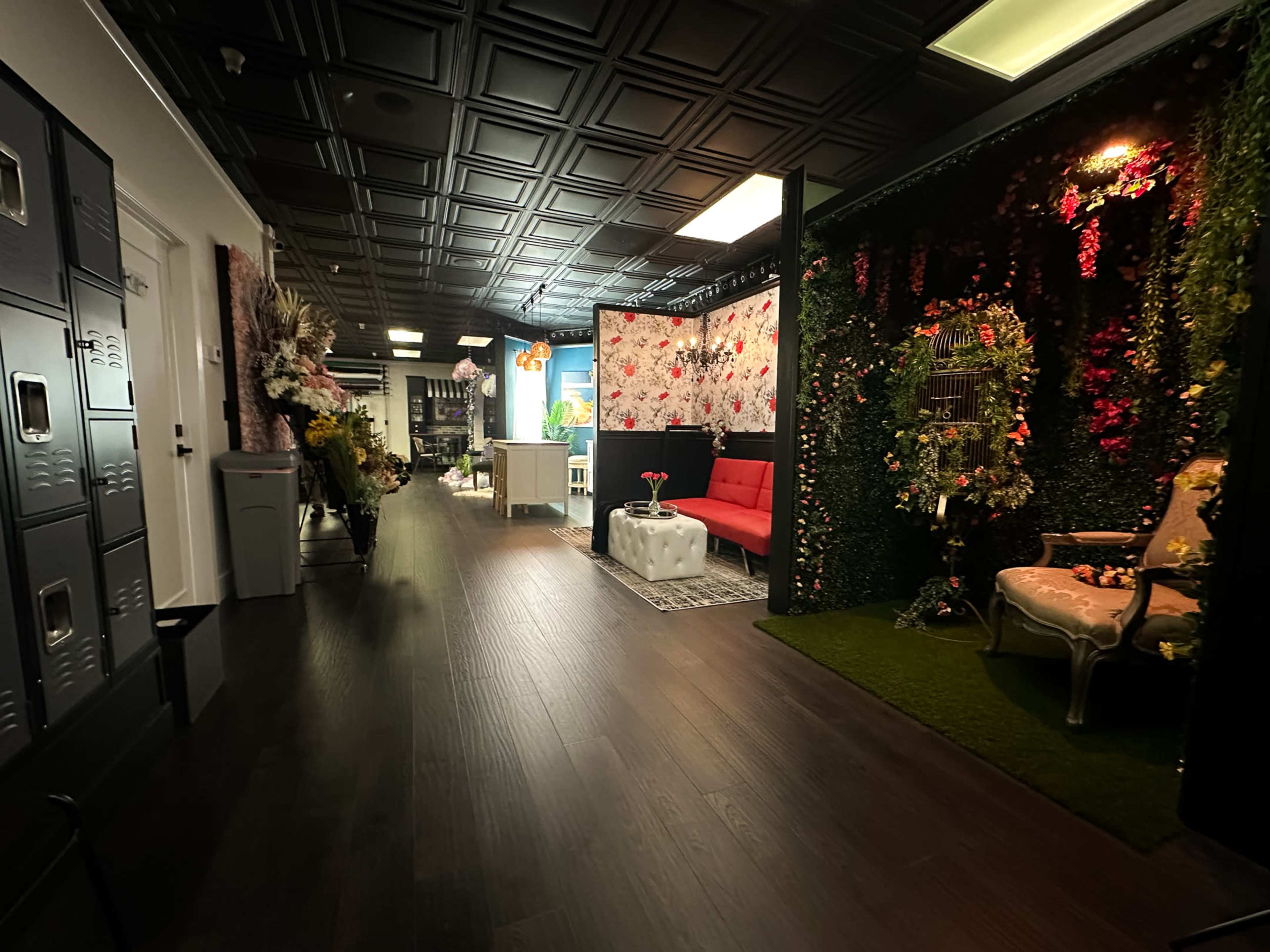 The image shows a hallway with decorative walls on one side, a red couch, lockers, and an area featuring lush greenery and floral accents.