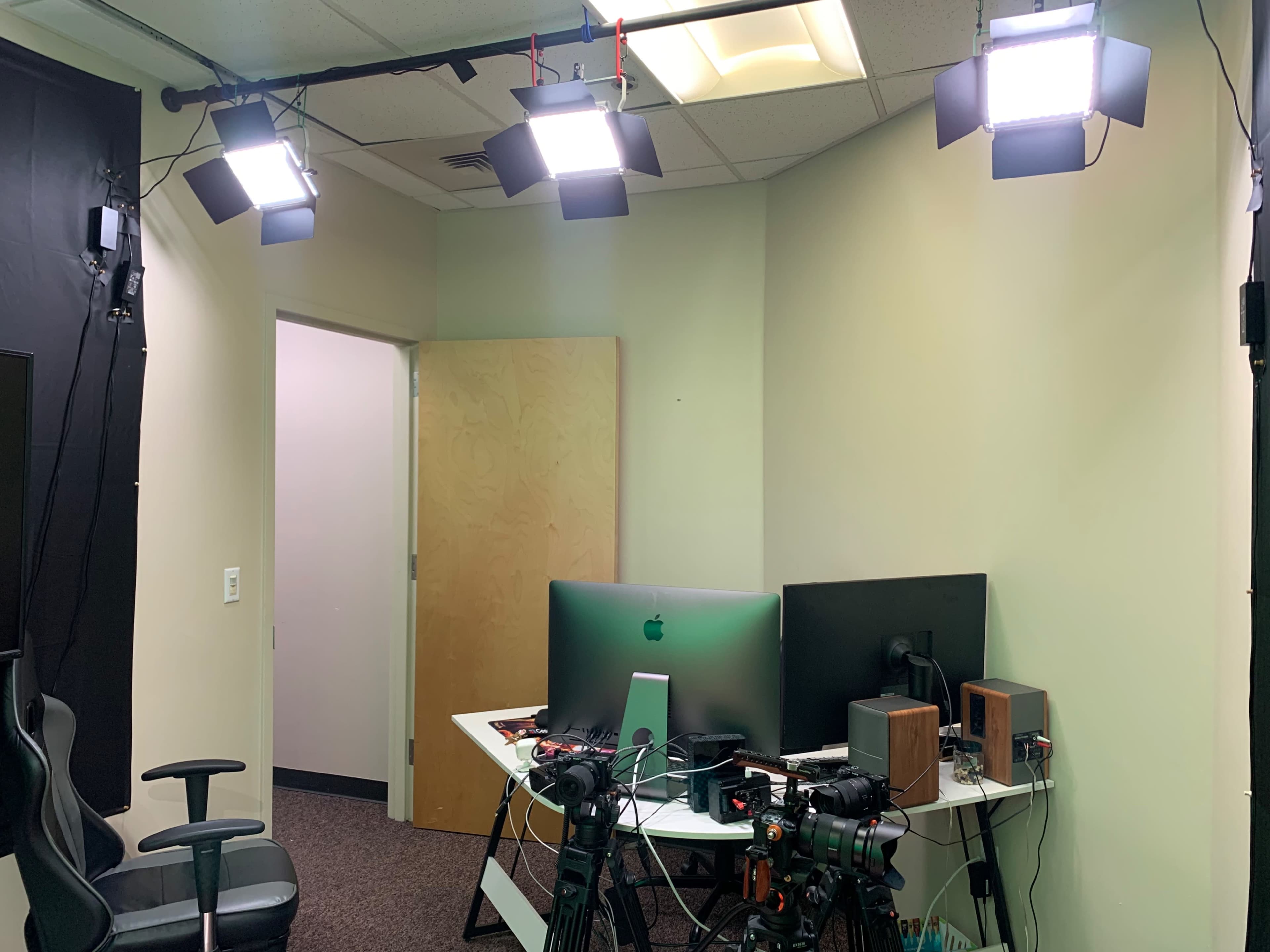 A dimly lit room with three ceiling lights illuminating a desk containing two computer monitors and cameras, while a door is visible in the background.
