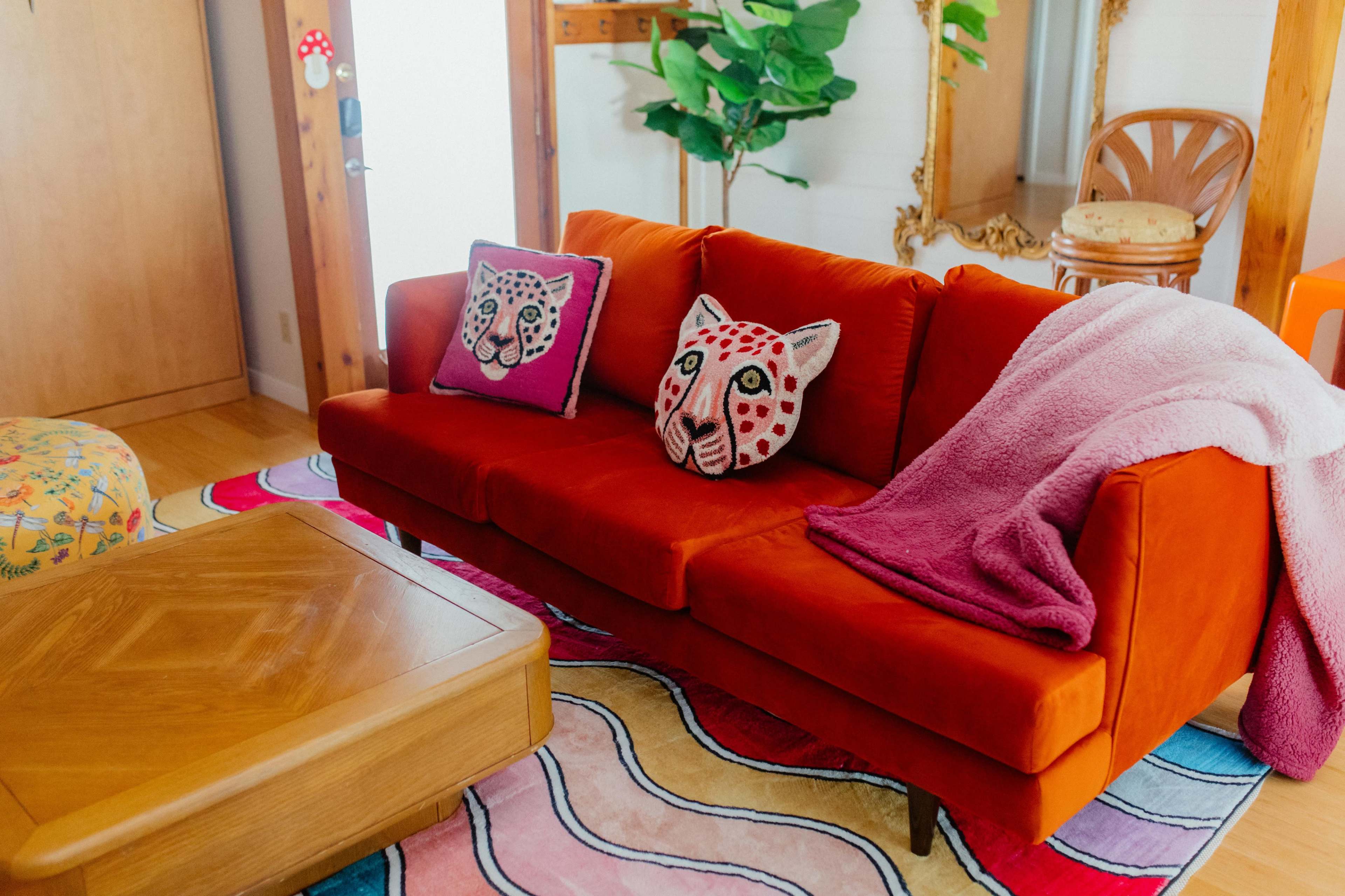 A bright orange sofa with patterned pillows and a purple throw blanket is positioned on a colorful rug in a cozy living room.