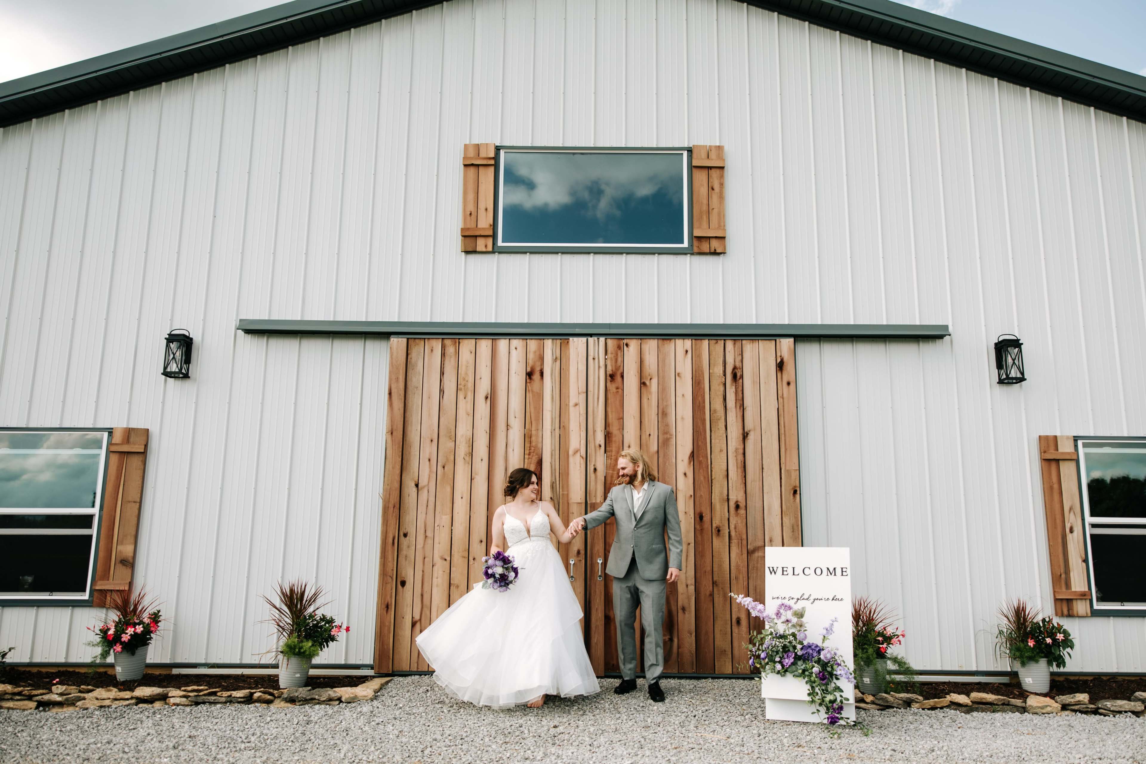 A bride and groom walk hand in hand in front of a modern barn venue adorned with wooden accents and flower arrangements.