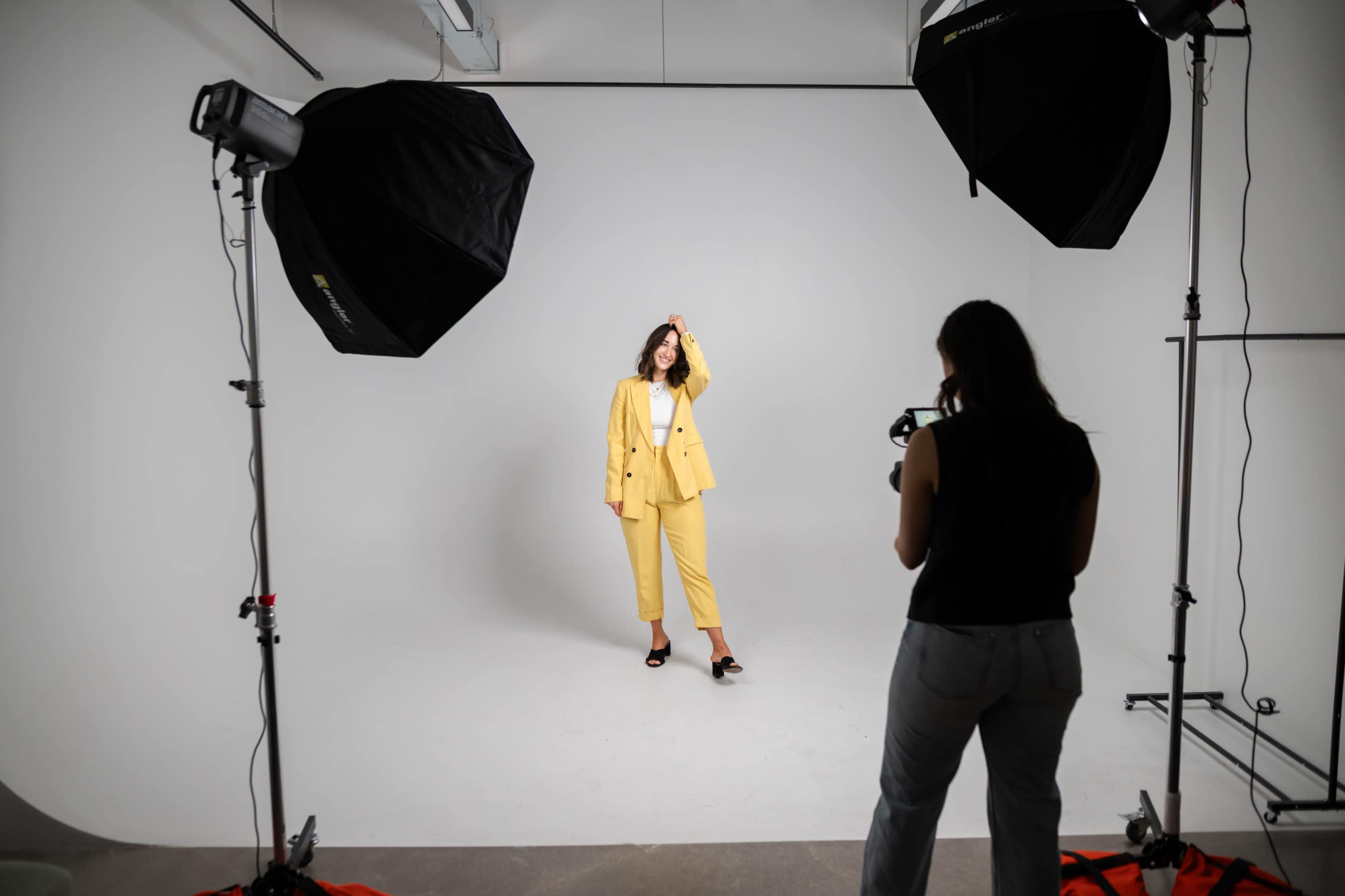 A woman in a yellow suit poses in a studio while another woman takes her picture with a camera.