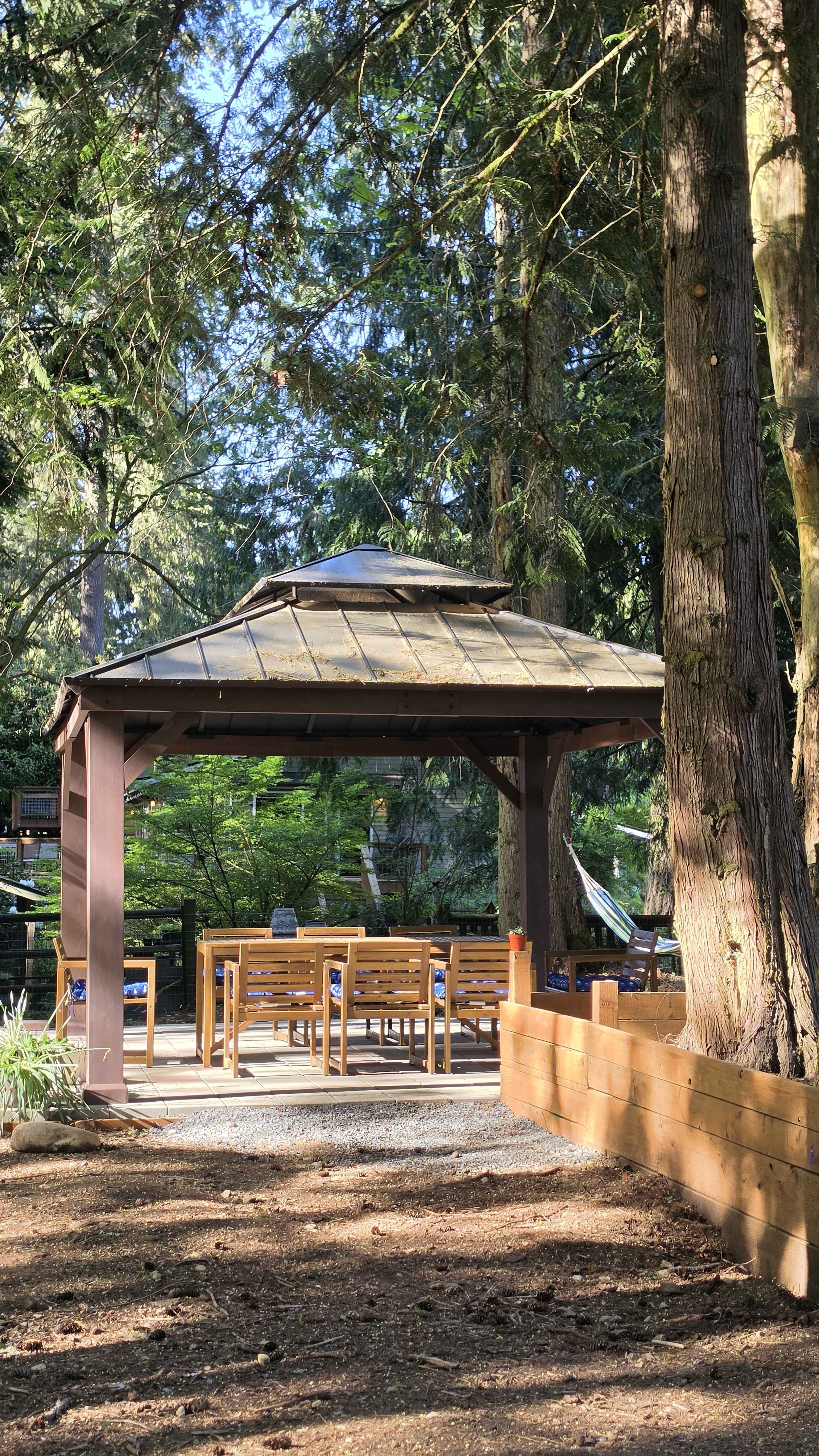 The image shows a wooden gazebo surrounded by tall trees, featuring a set of tables and benches inside.