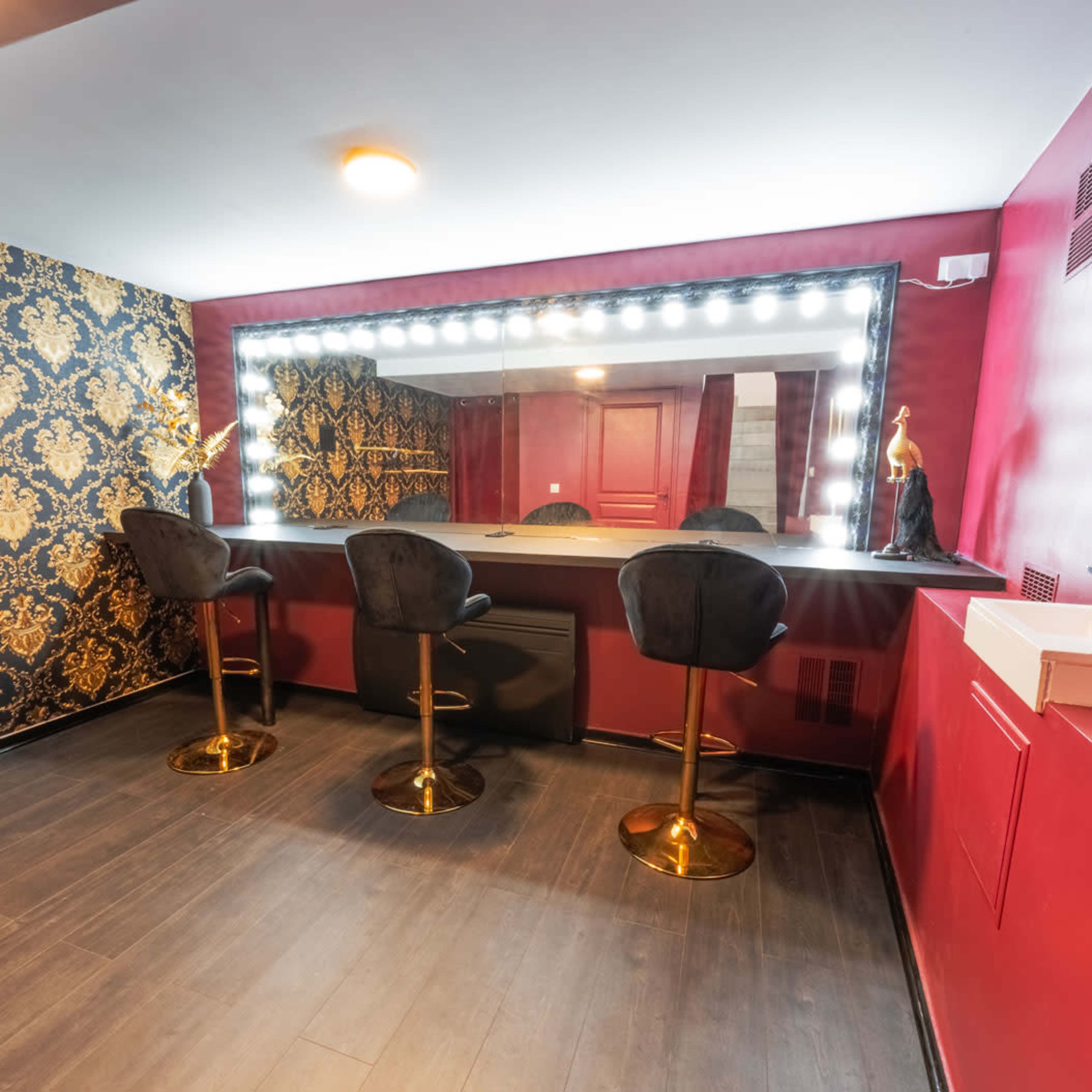 The image shows a chic vanity area with three black bar stools, a large illuminated mirror, and a decorative wall featuring a gold and black patterned design.