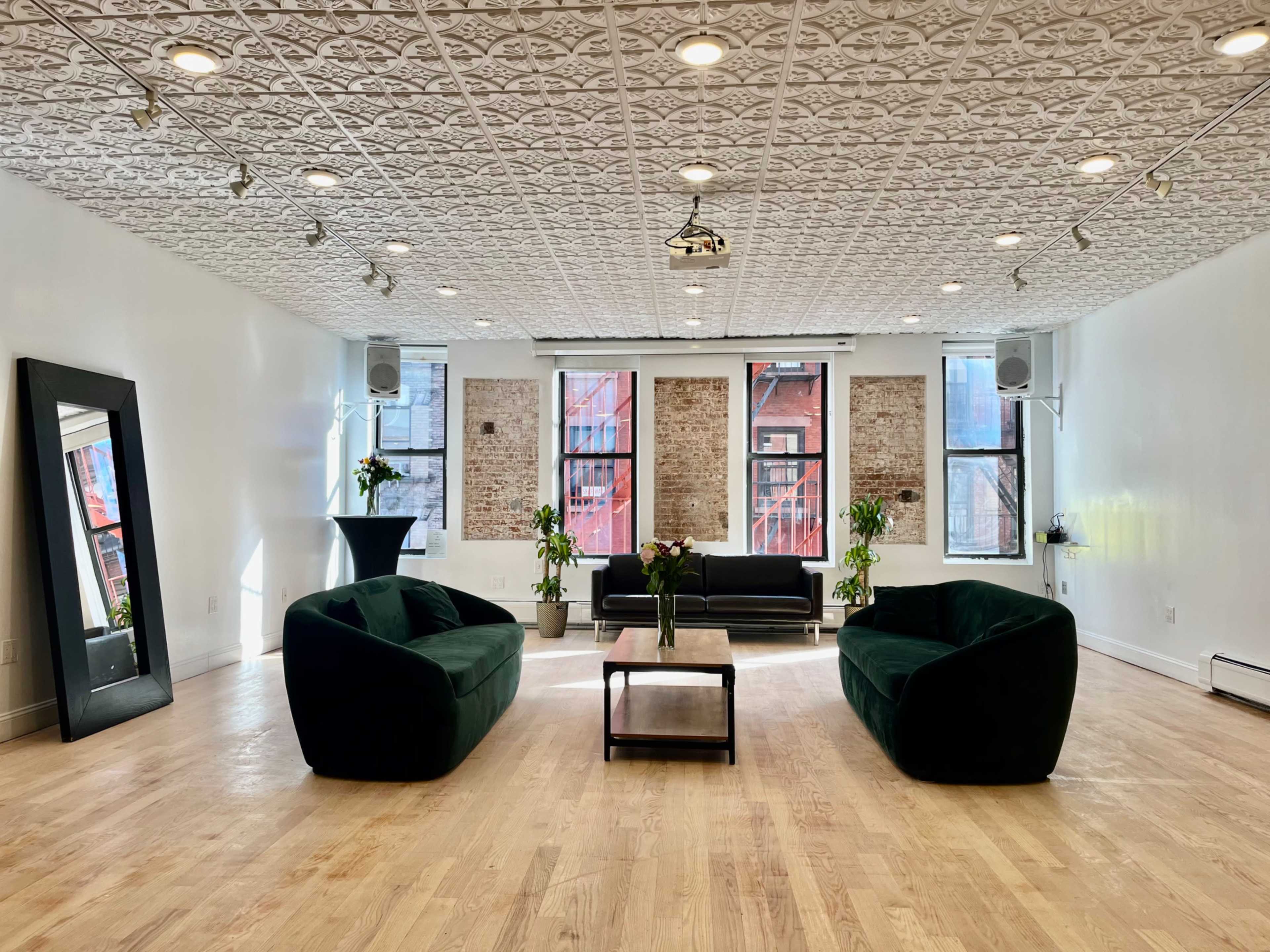 The room features two green sofas, a black couch, a coffee table, and a mirror, all set against a backdrop of exposed brick walls and bright windows.