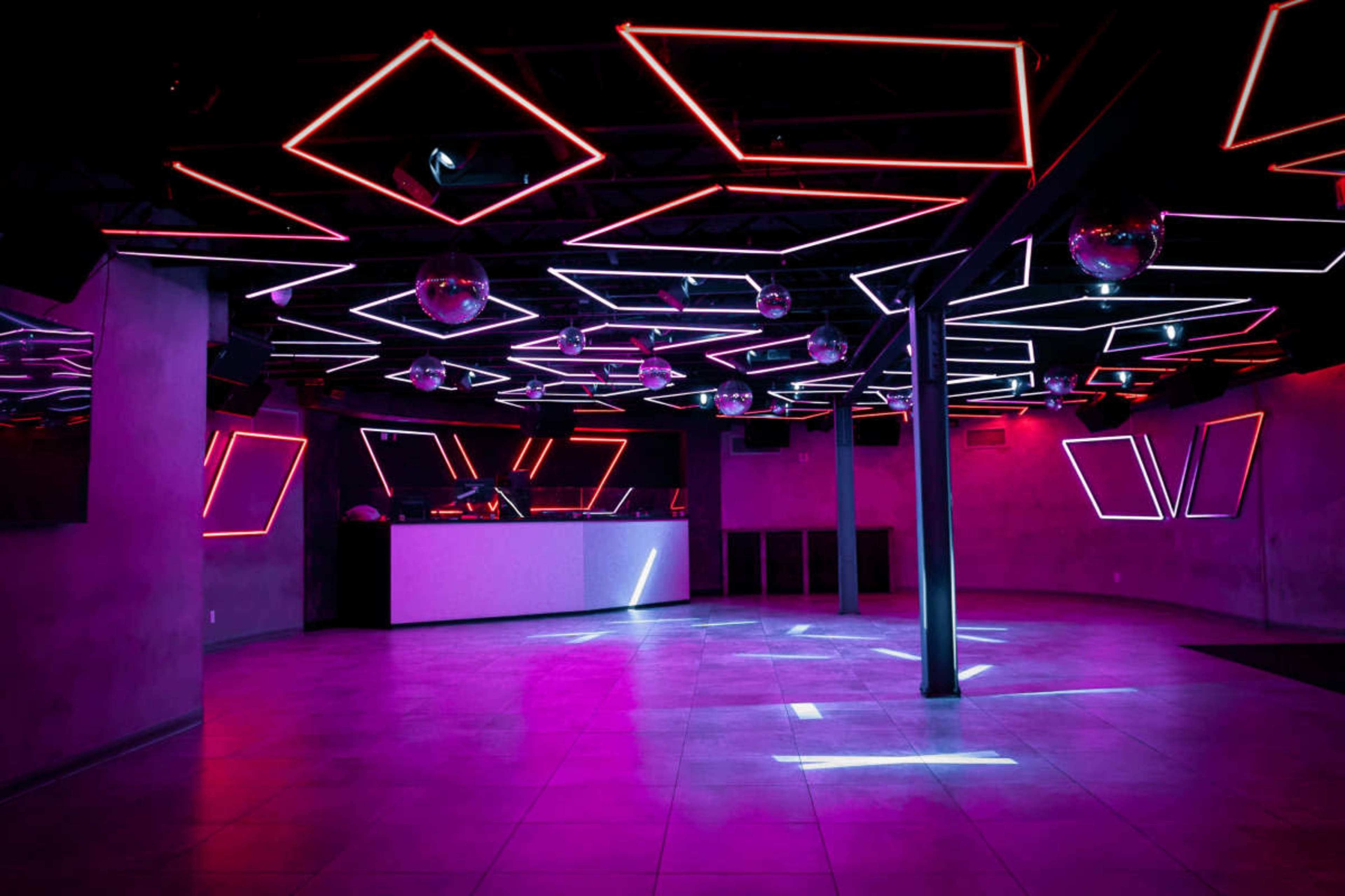 The image displays a dimly lit nightclub featuring geometric neon lights on the ceiling and a bar area in the background.
