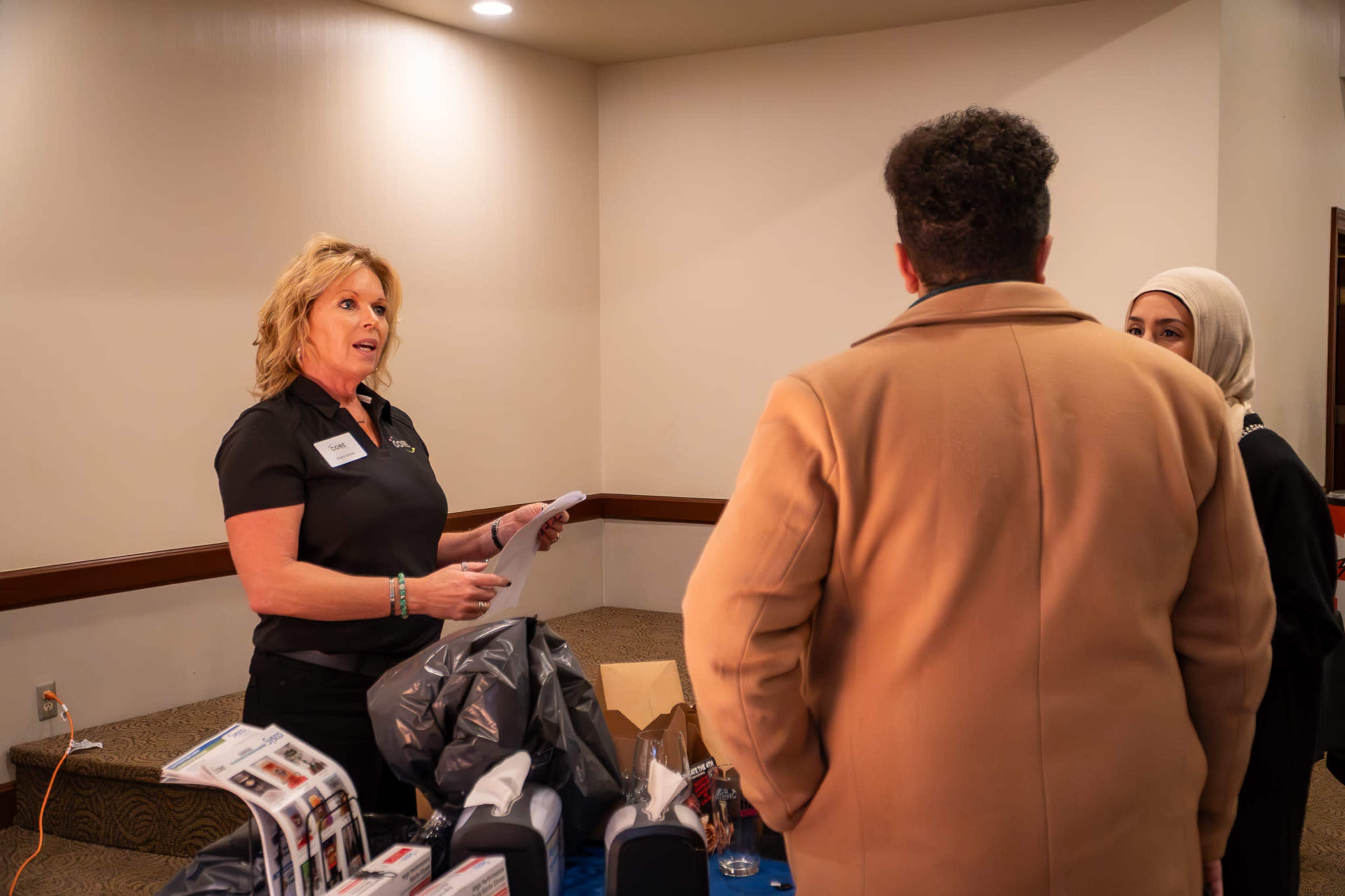 A woman in a black shirt discusses information with two individuals at a table displaying promotional materials.