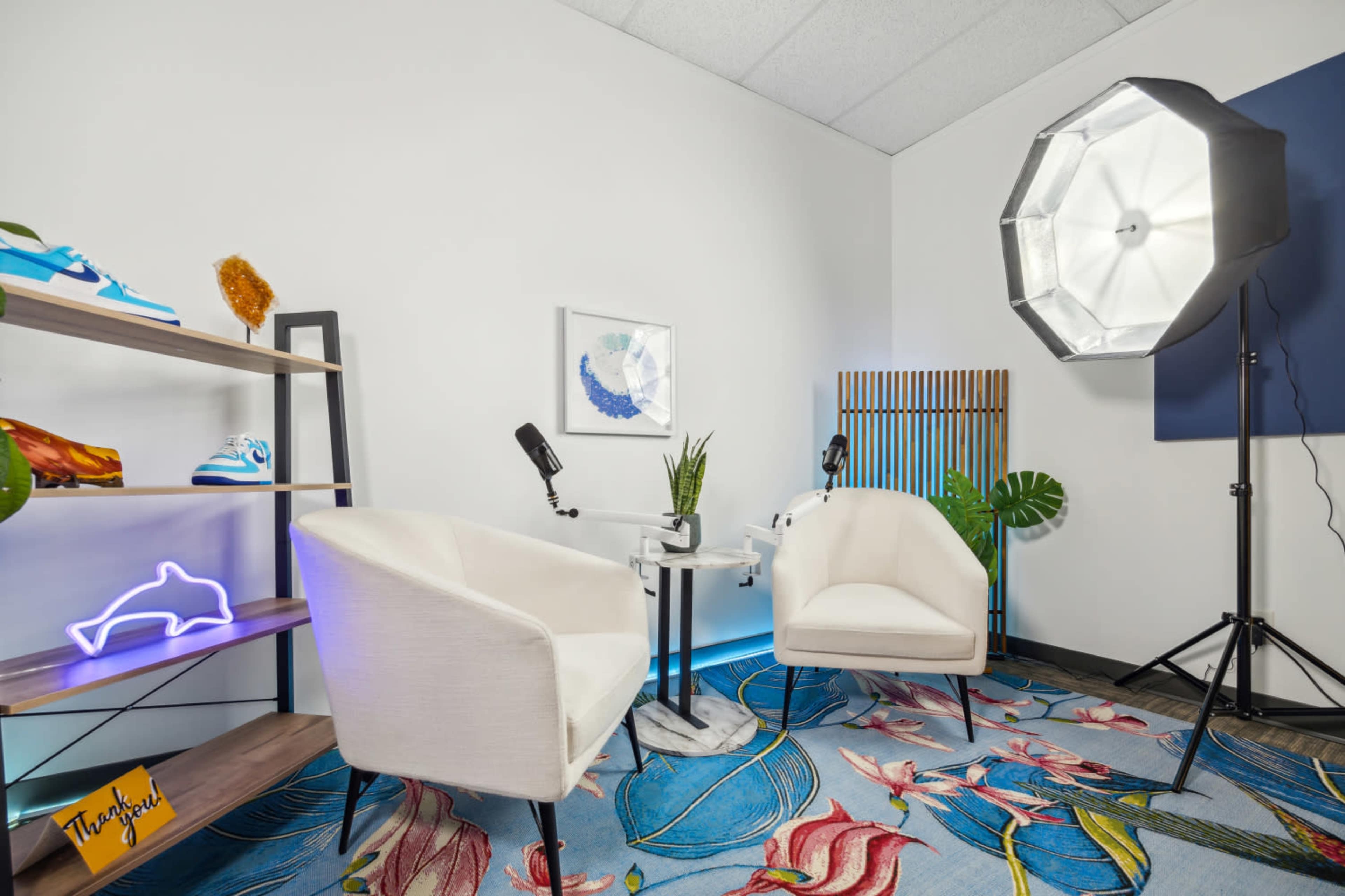 A small indoor space with two white chairs, a glass table, a decorative plant, a shelf displaying items including sneakers and a neon sign, and a large softbox light setup.