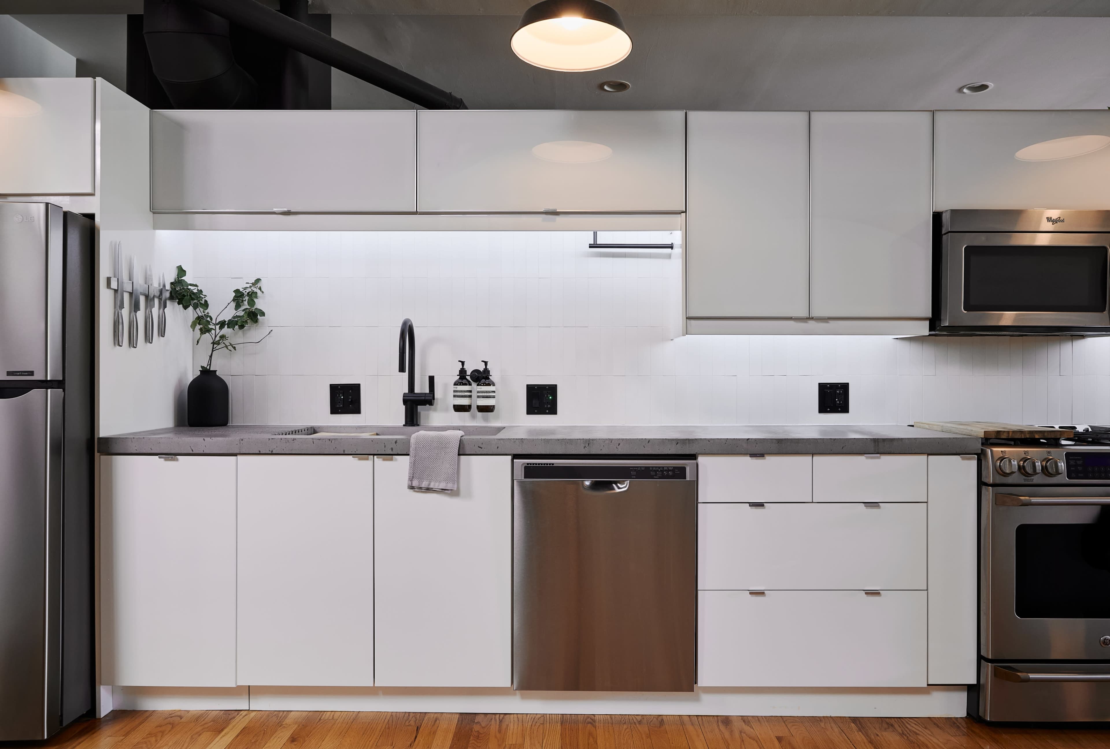 The image shows a modern kitchen featuring white cabinetry, stainless steel appliances, and a minimalist design with a concrete countertop.