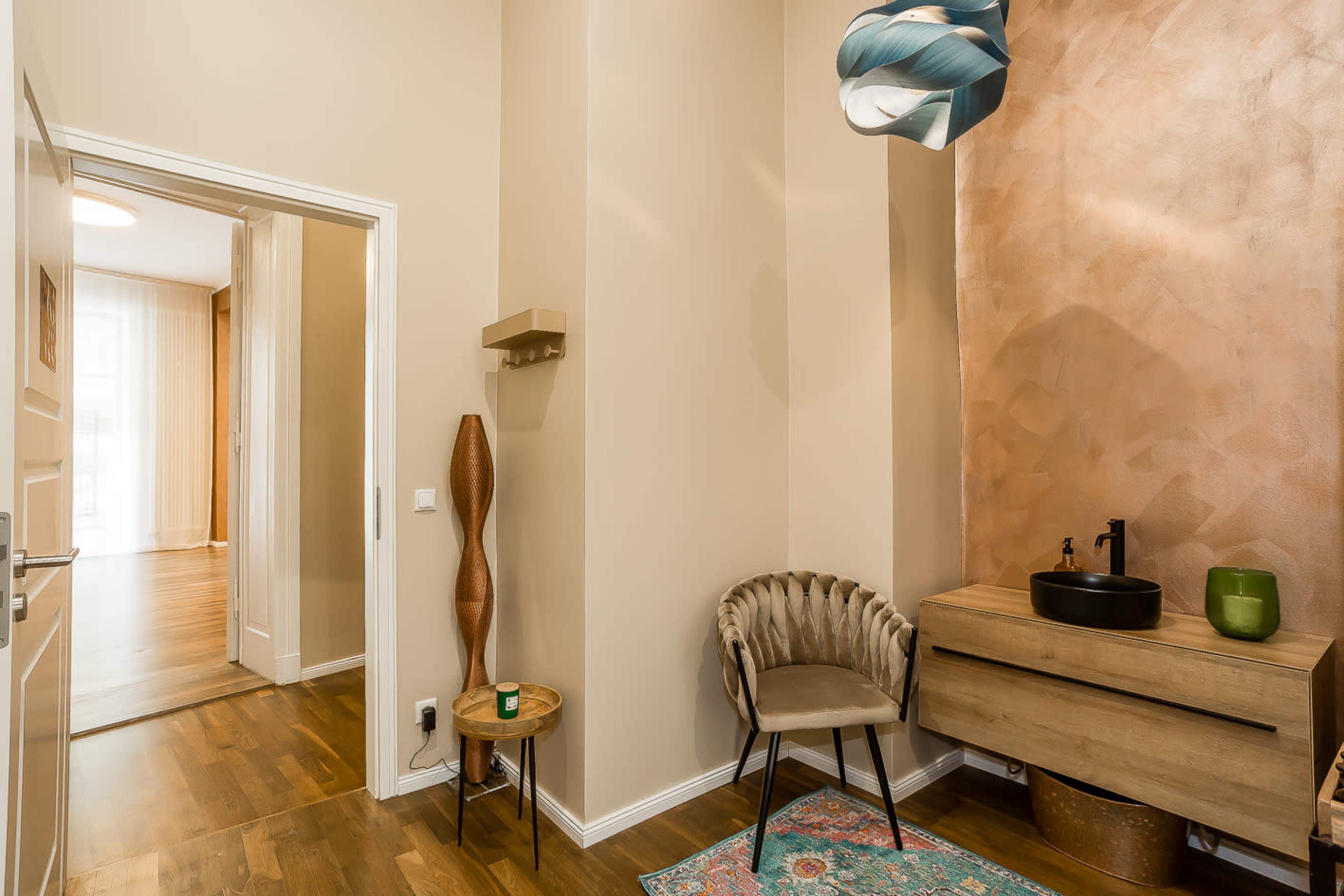 A modern hallway featuring a wooden console table, a round black sink, a stylish chair, a decorative vase, and a textured wall with warm tones.