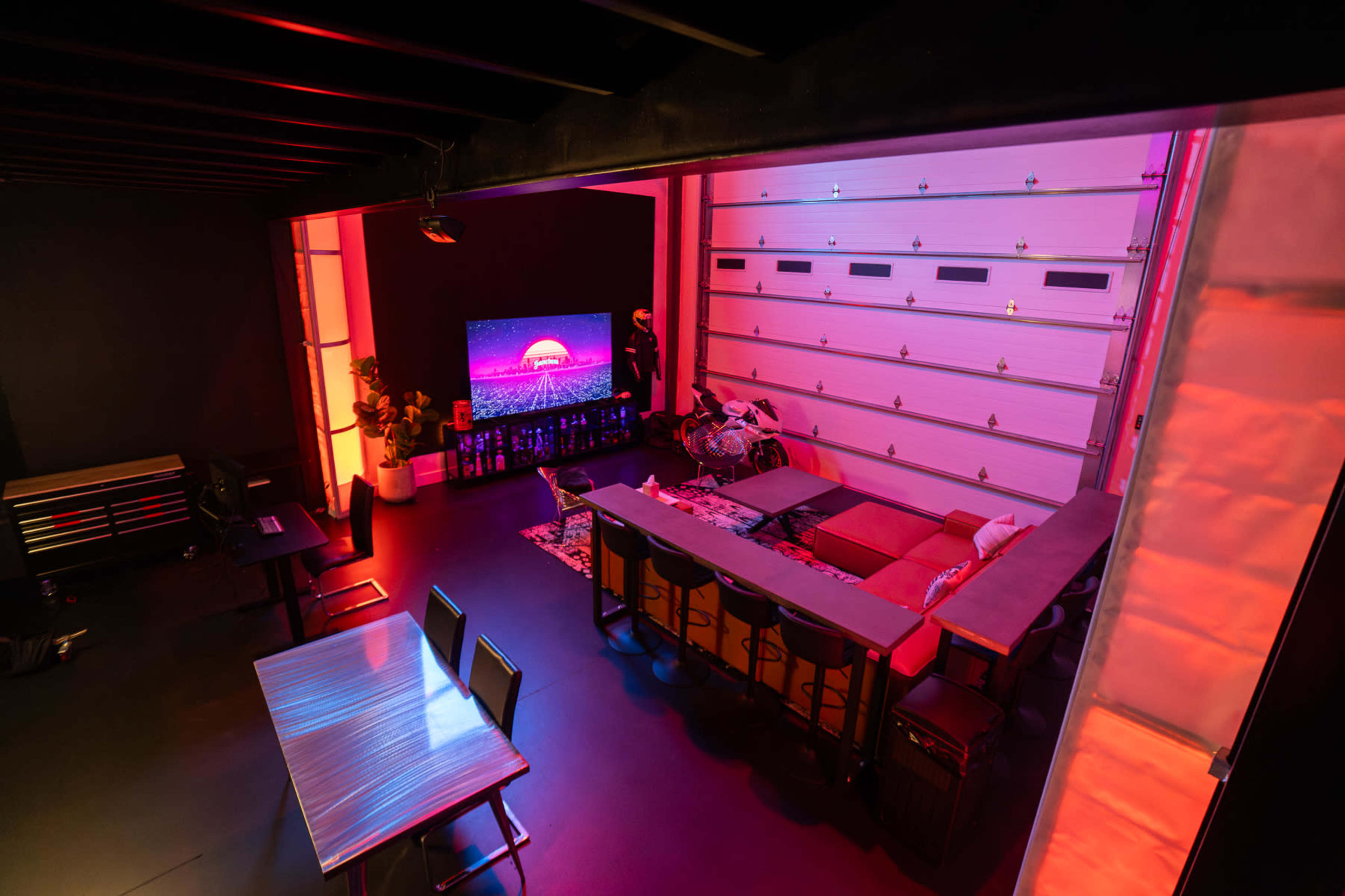 The image shows a modern indoor space with a large screen, seating area, and a dining table, illuminated with colorful lighting.