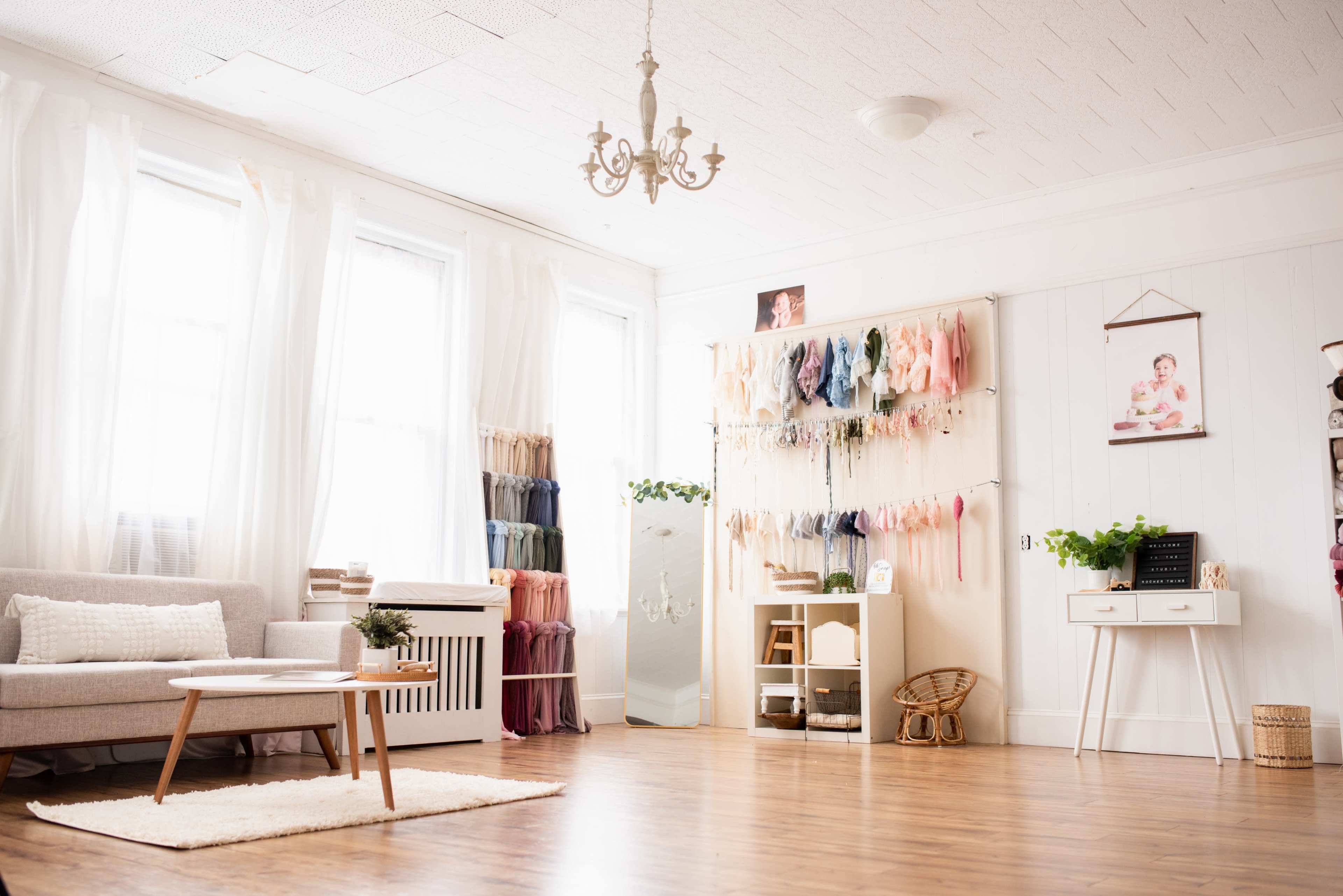 A bright, airy room features a sofa, wooden coffee table, decorative shelves filled with colorful fabric swatches, and a wall with framed photographs and indoor plants.