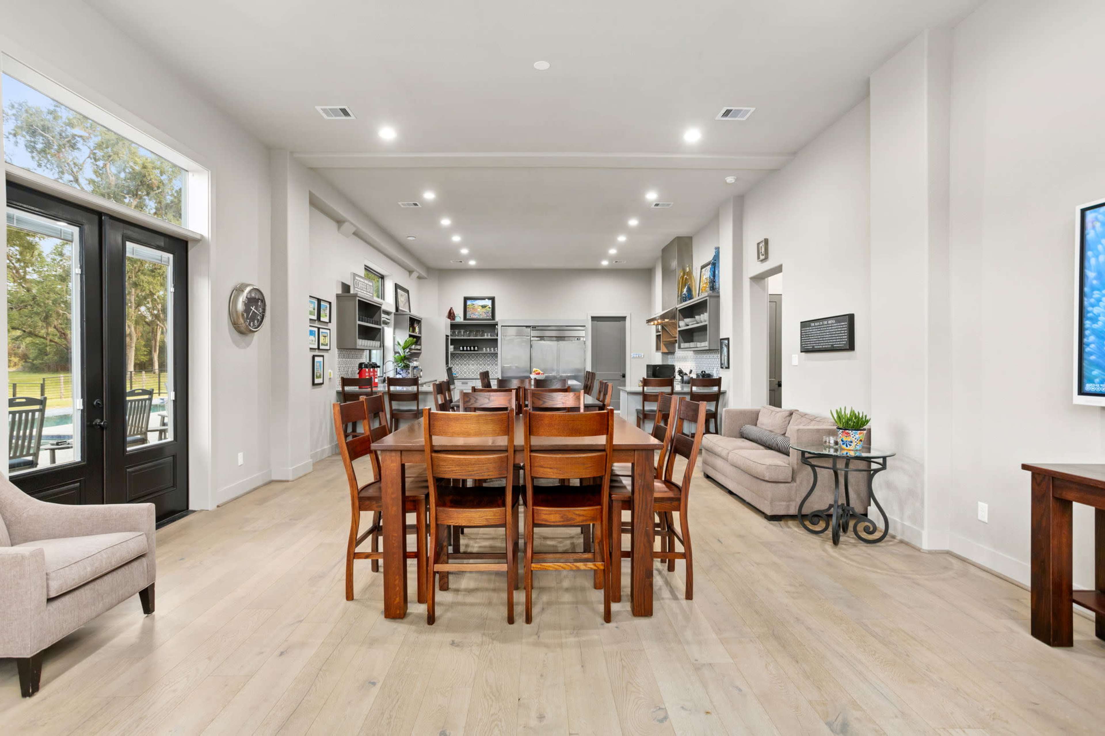 A spacious room with a large wooden dining table surrounded by chairs, adjacent to a seating area and kitchen appliances along the back wall.