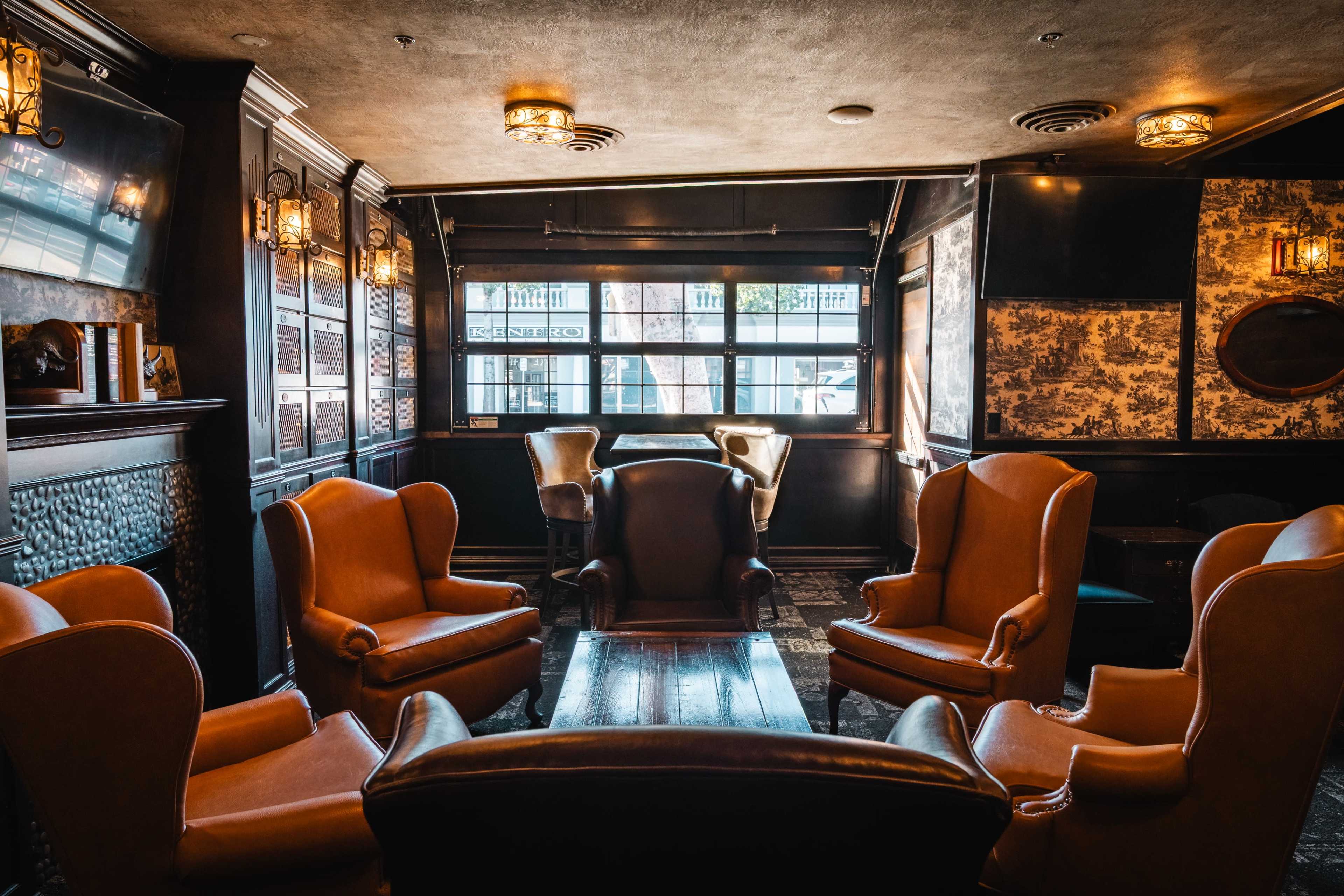 The image shows a cozy, well-decorated lounge area with brown leather wingback chairs arranged around a wooden table, and large windows letting in natural light.
