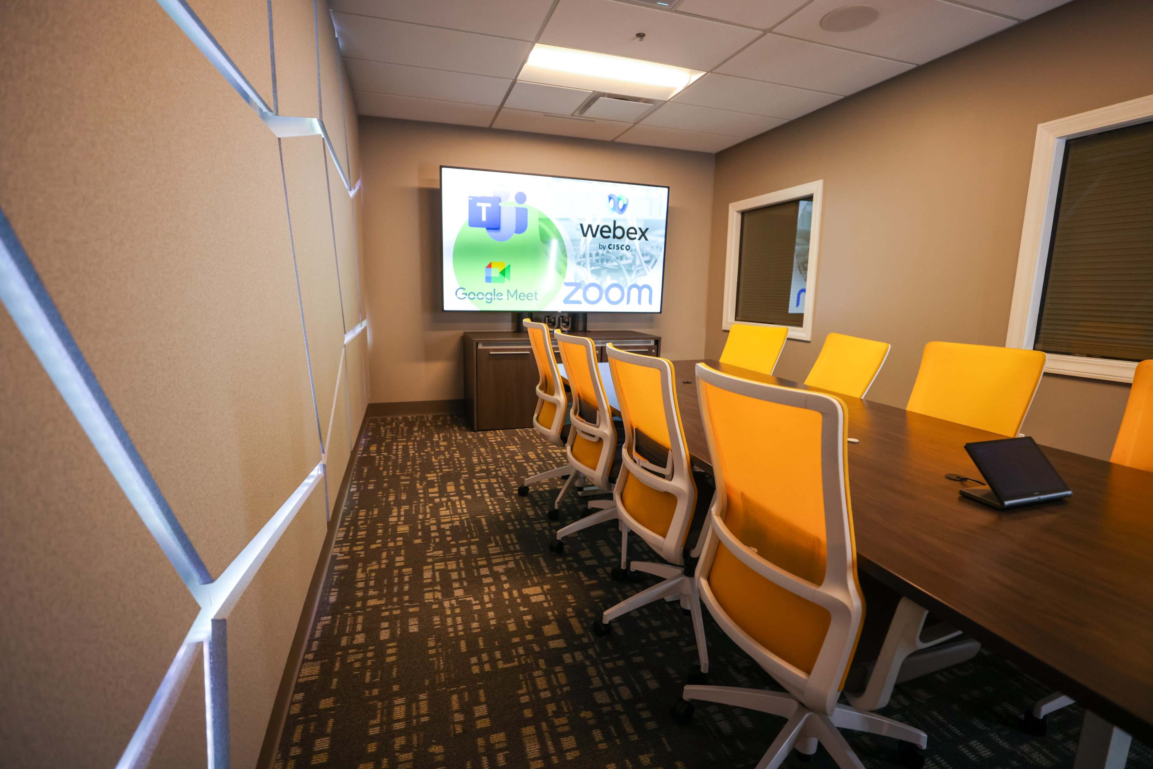A modern conference room with a large screen displaying various video conferencing logos and a long table surrounded by orange chairs.