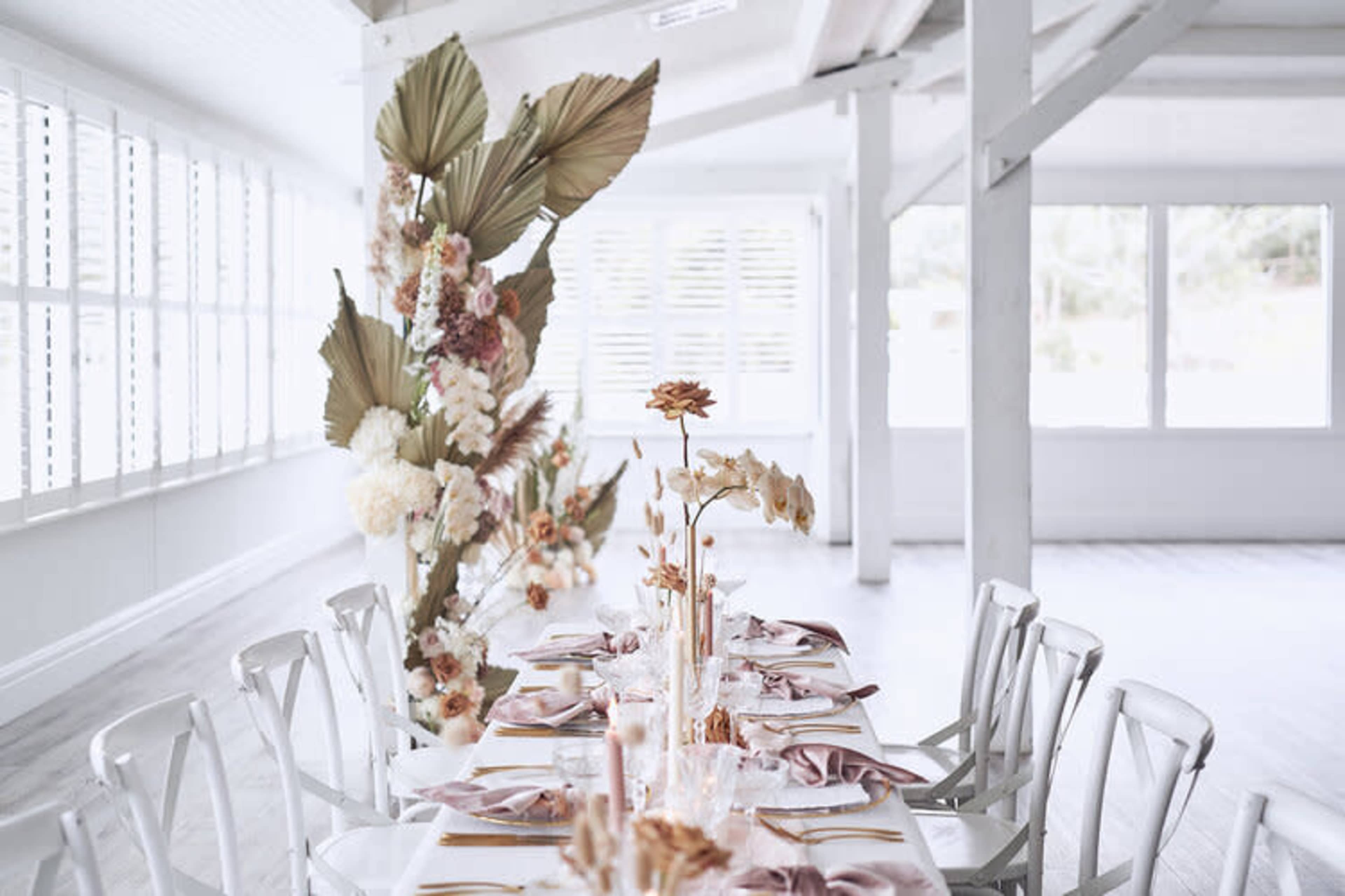 A long dining table is elegantly set with decorative flowers and napkins in a brightly lit, minimalist room.