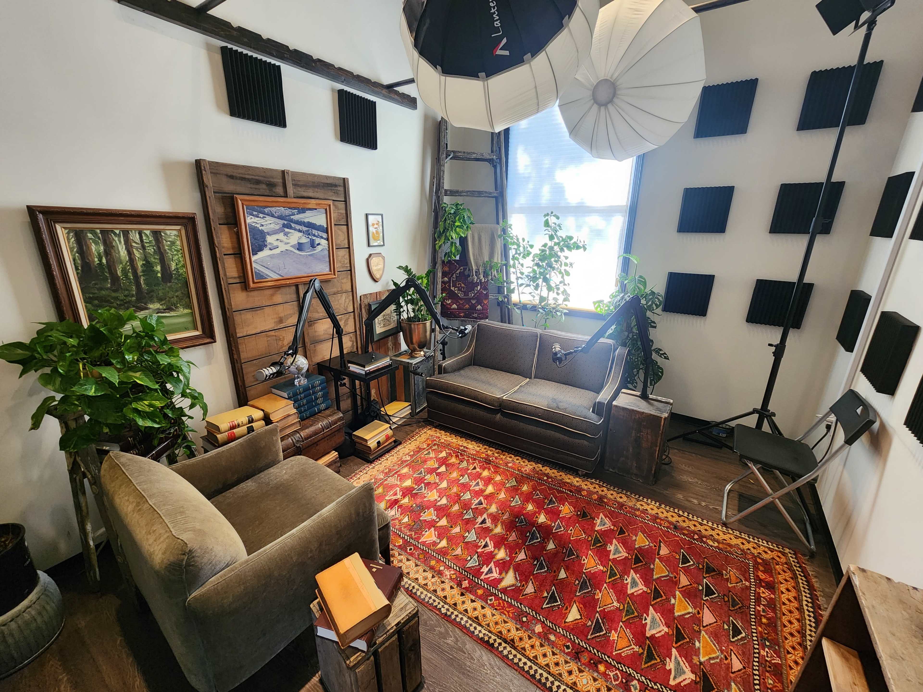 The image shows a cozy interior room with a patterned rug, two couches, a bookshelf, potted plants, and various photographic lighting equipment set up.