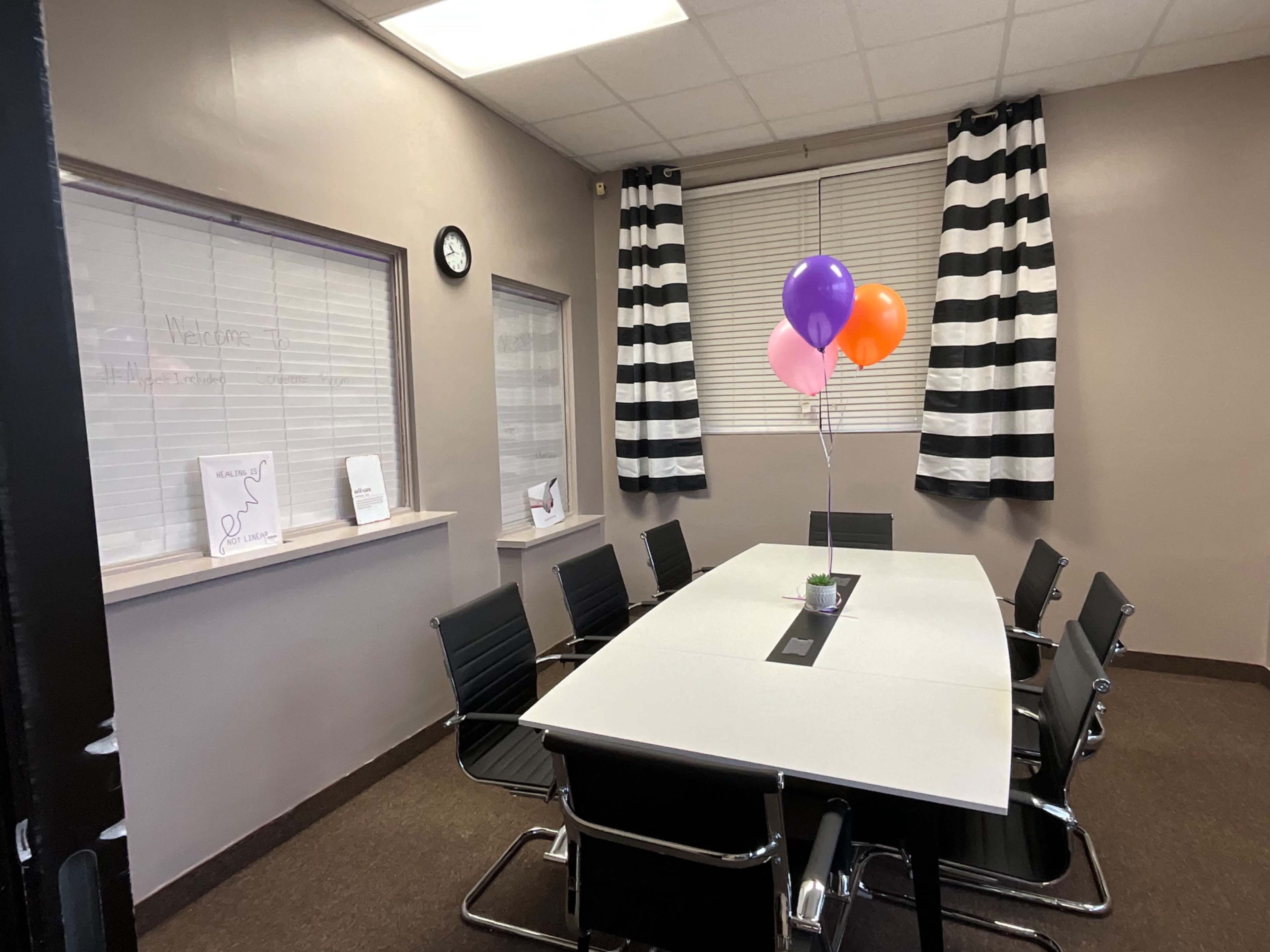 A conference room with a long white table, black chairs, and a small plant with three balloons on it, framed by striped curtains.