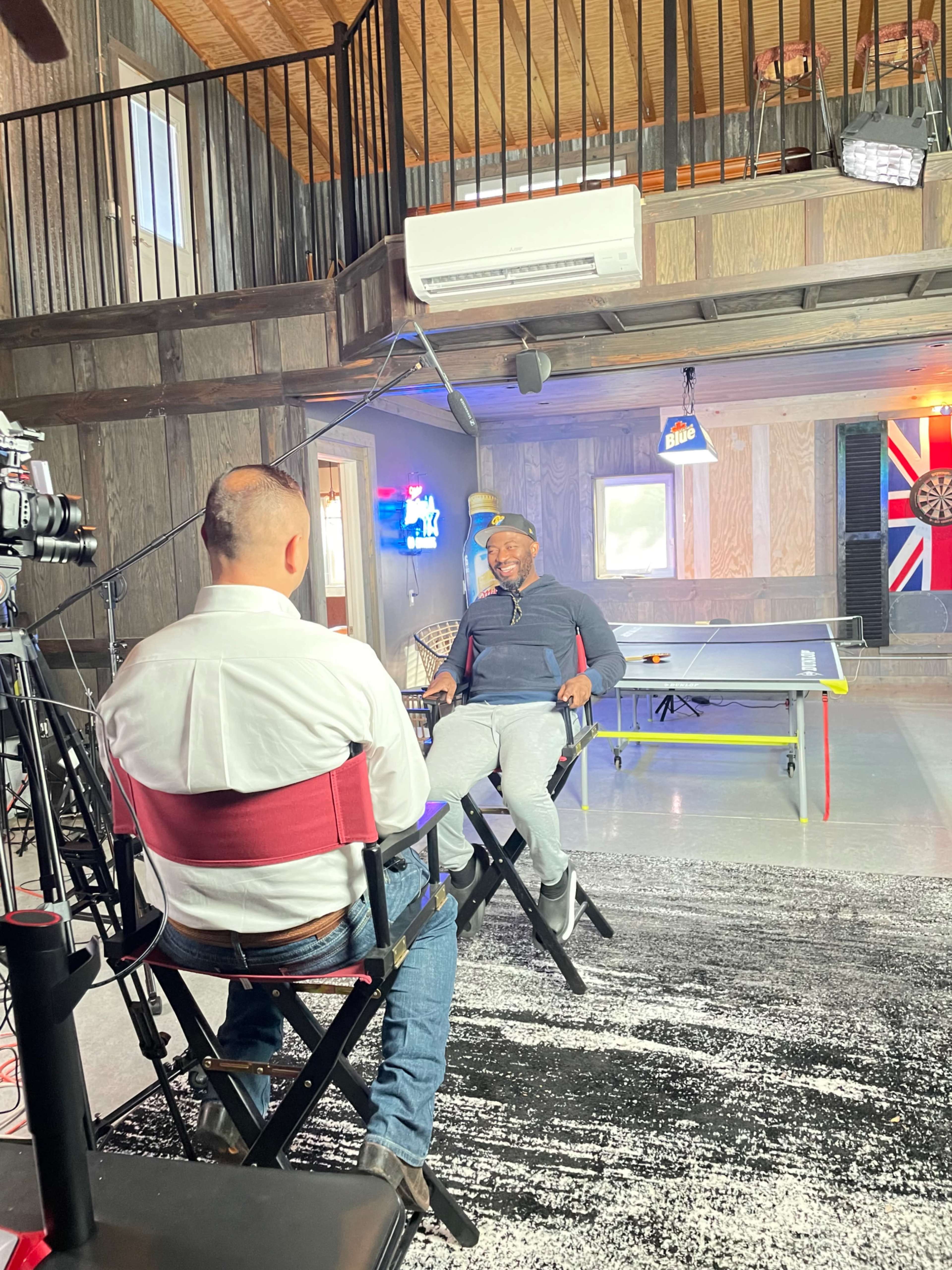 A person is being interviewed in a well-lit room with wooden walls and a ping-pong table in the background.