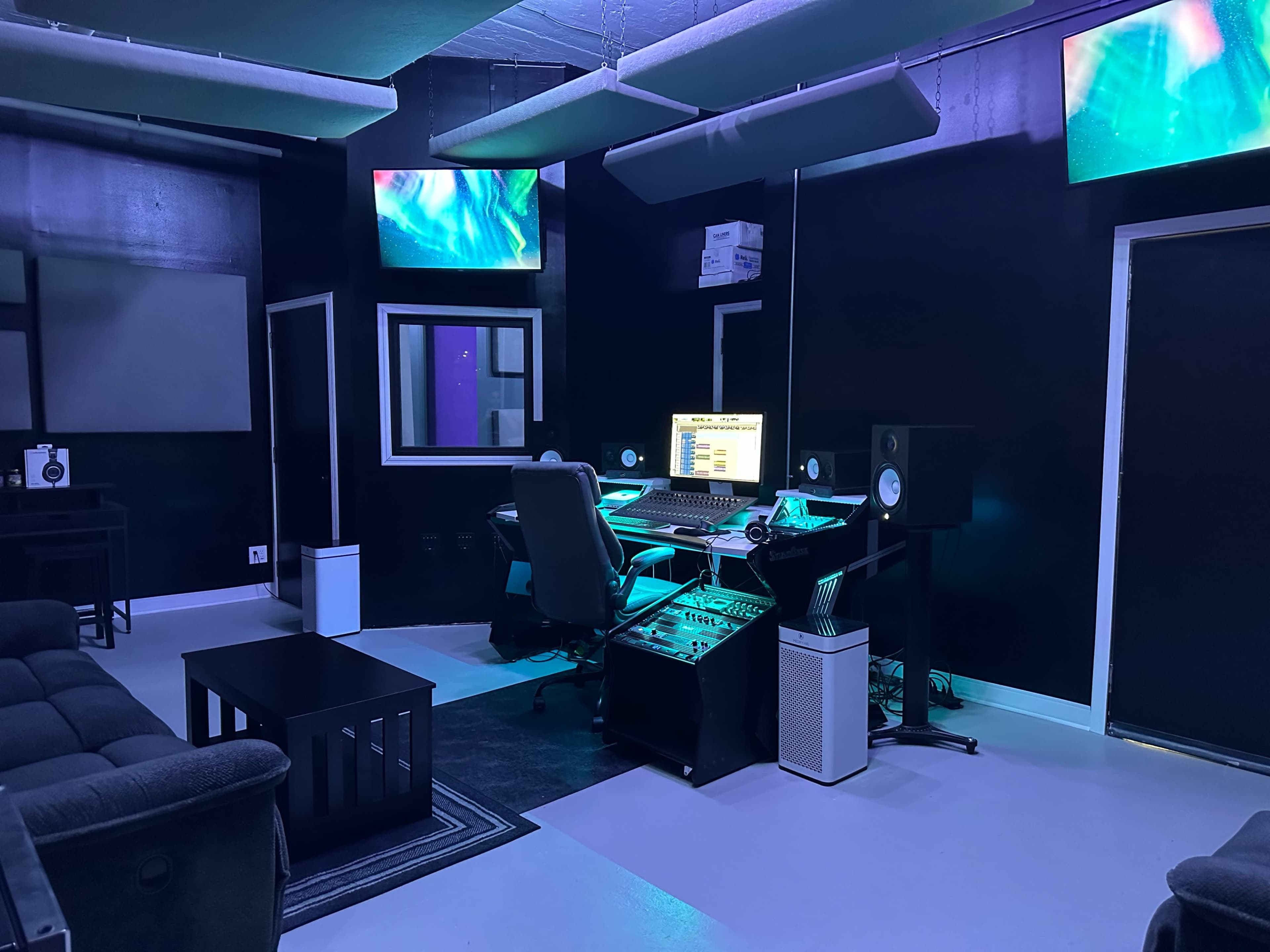 A music studio features a desk with a computer, audio equipment, and sound monitors, surrounded by dark walls and illuminated screens.