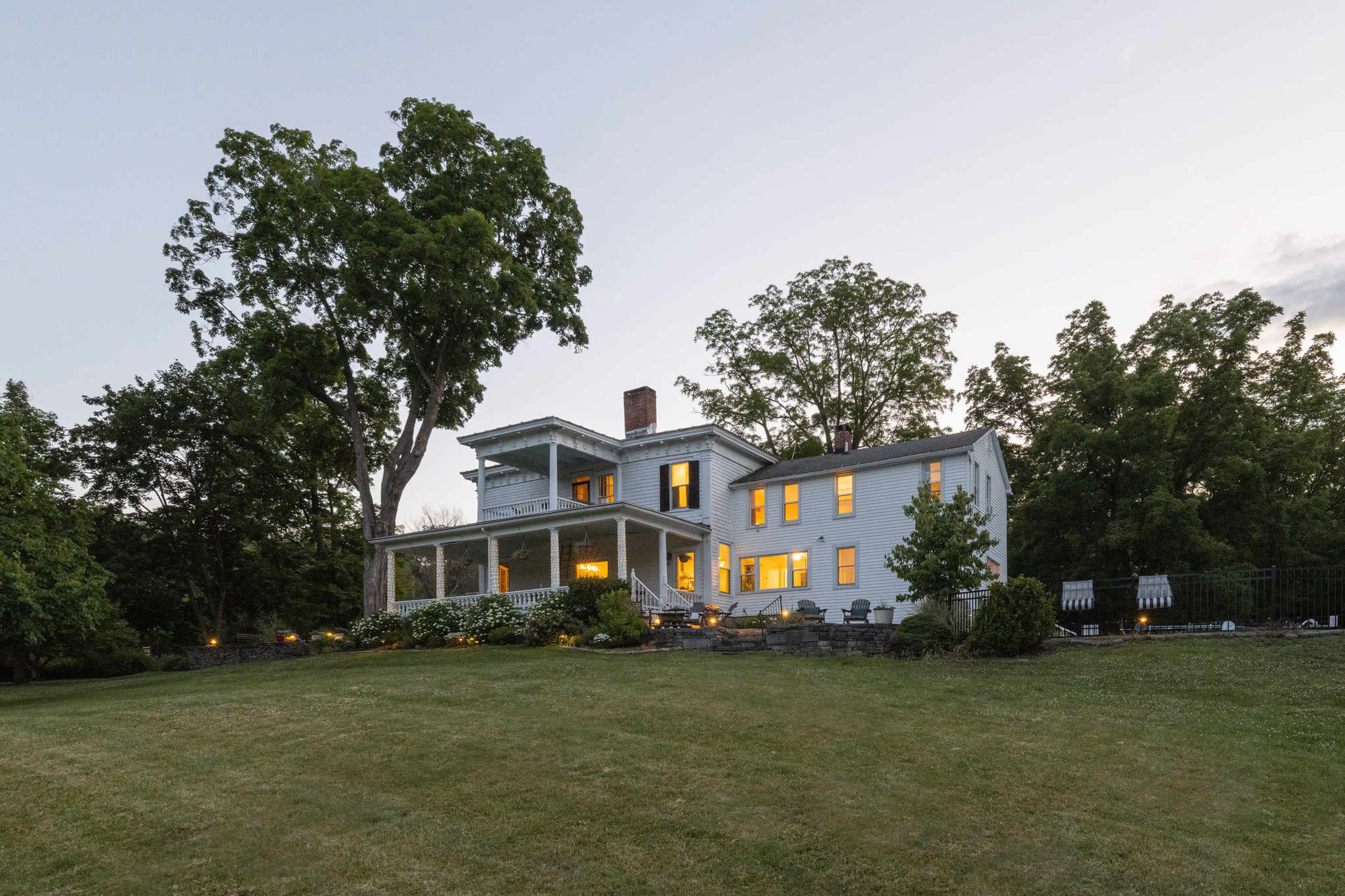 A large, two-story house with multiple windows is set against a backdrop of trees in a green yard during twilight.