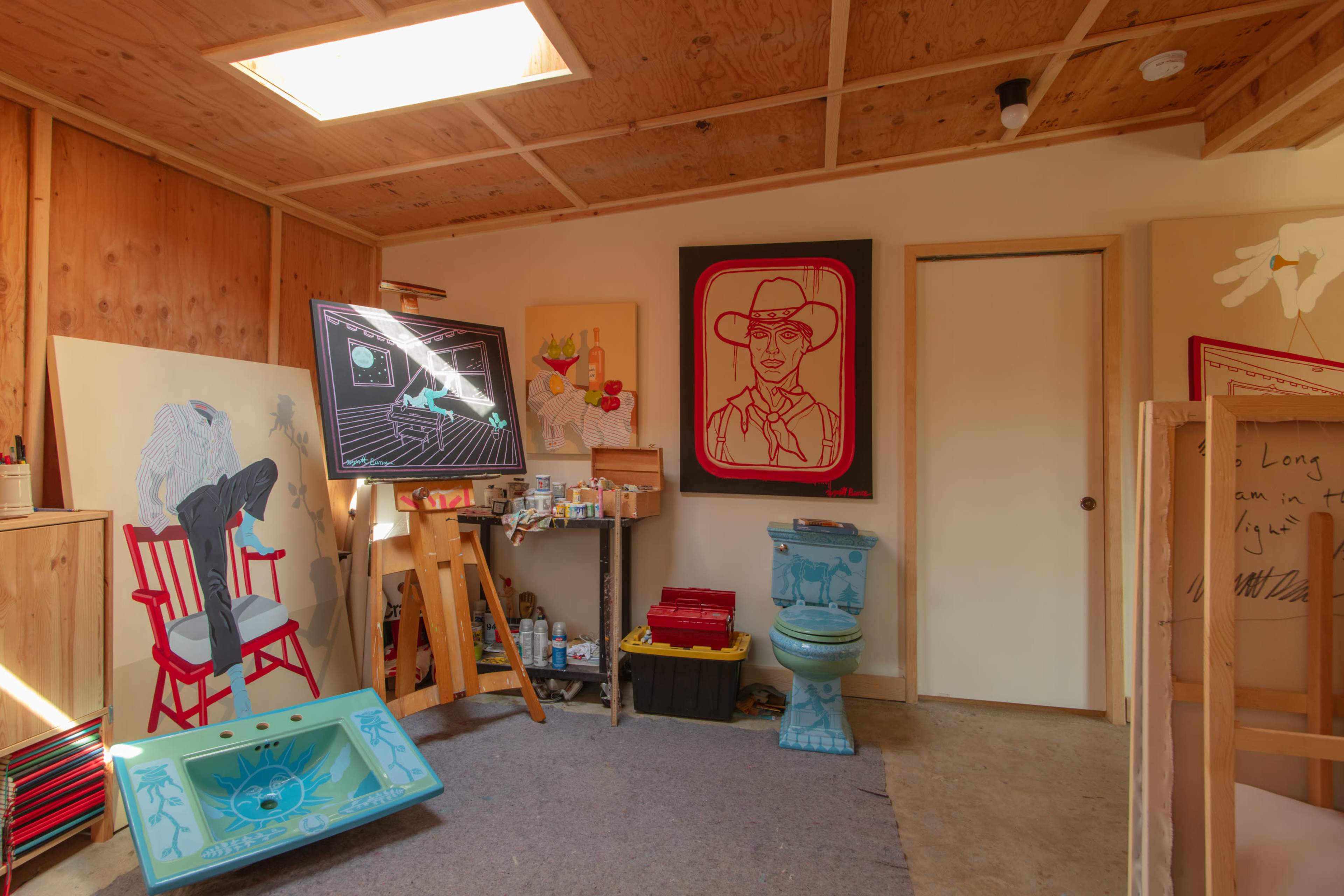 The image shows an artist's studio with various artworks displayed on the walls, an easel with a painting, and a colorful toilet used as a decorative piece.