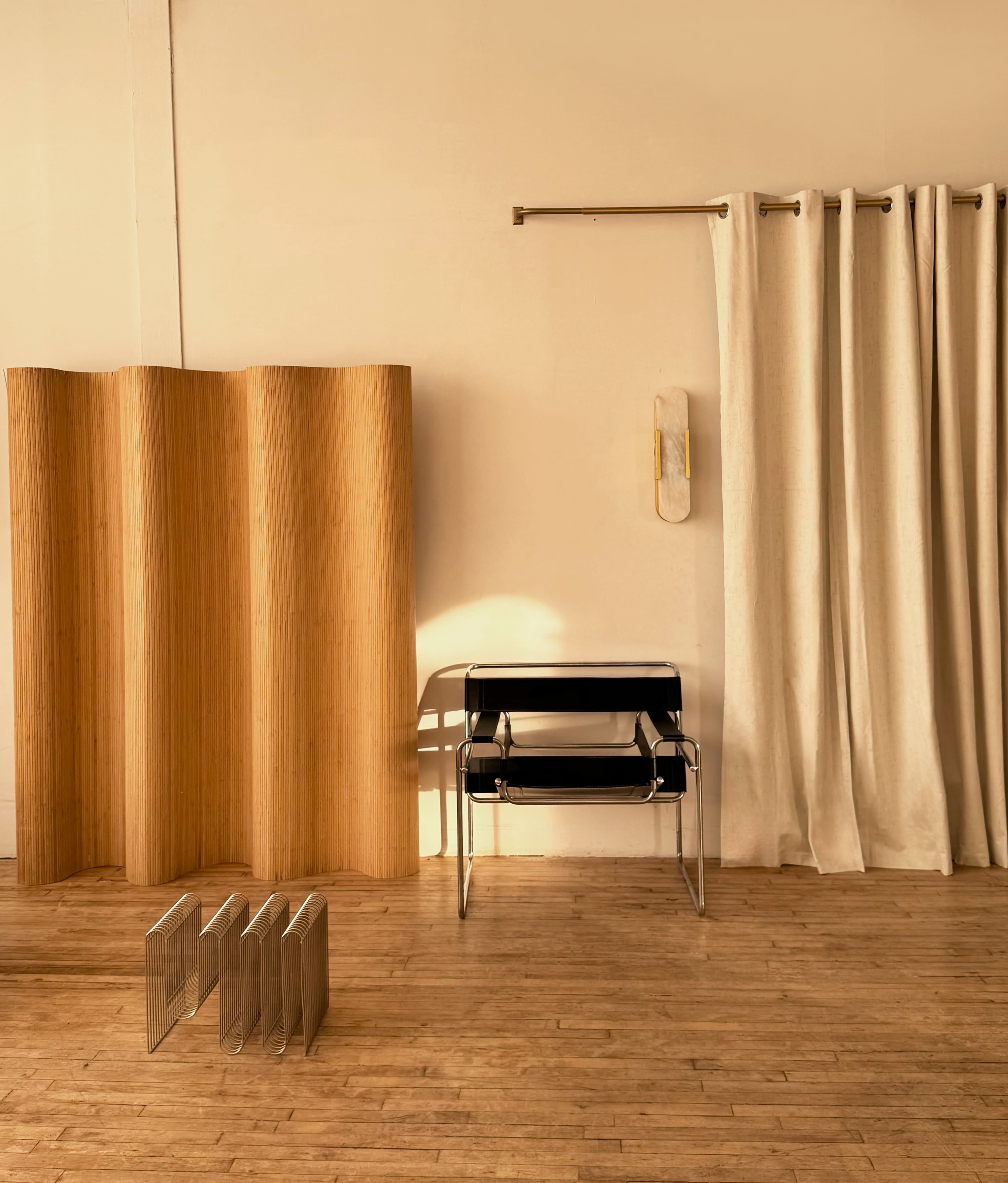 The scene features a minimalist interior with a wooden room divider, two black chairs stacked on top of each other, and light-colored curtains against a plain wall.