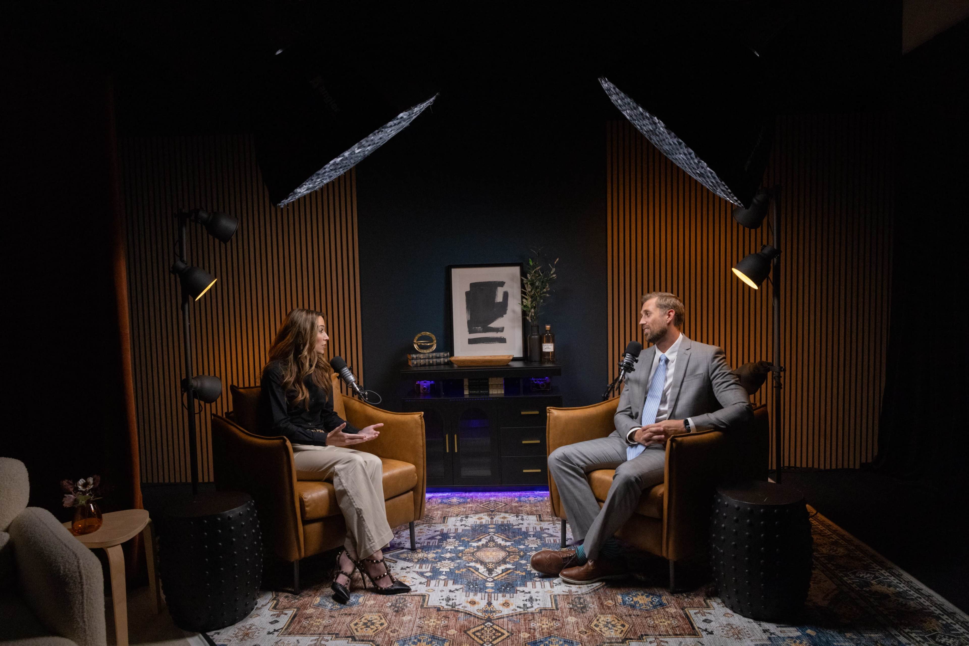 A woman and a man sit in leather armchairs facing each other in a well-lit interview set, with a decorative background featuring a framed artwork.