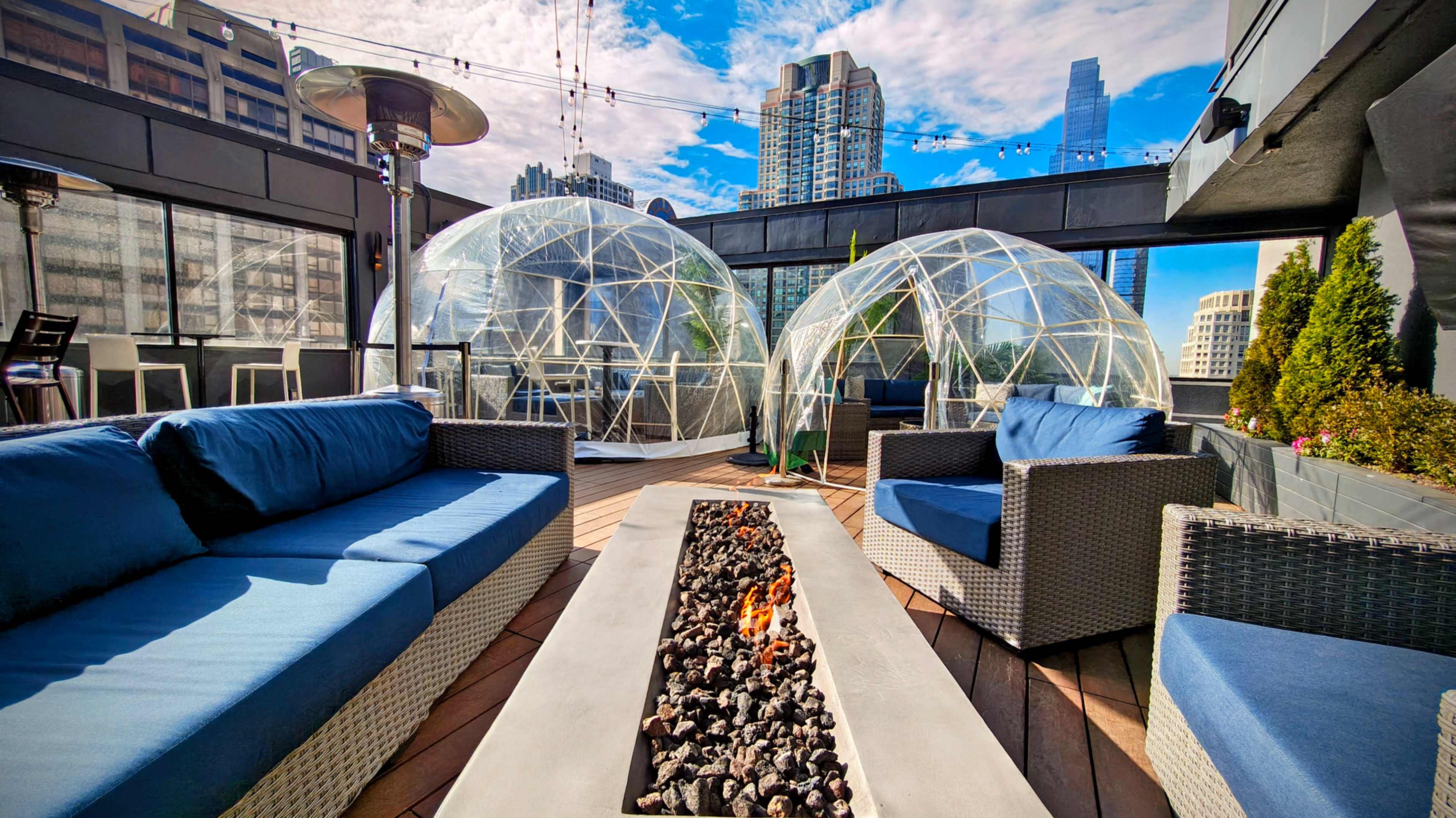 A rooftop lounge features modern seating arrangements and two transparent domes, with a fire pit set into a wooden deck surrounded by urban skyscrapers.