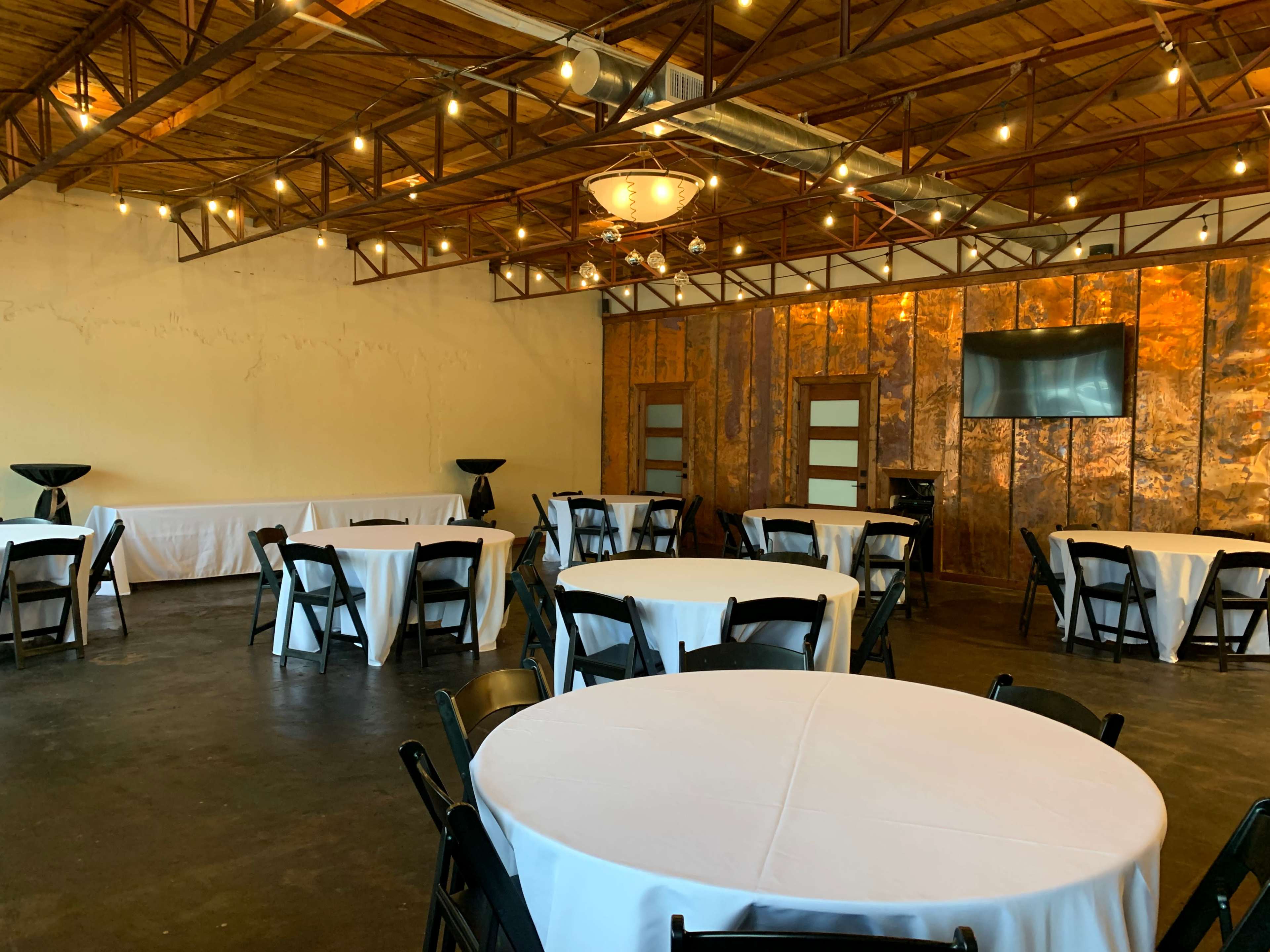 A spacious event room set up with round tables covered in white linens, black chairs, and exposed wooden beams overhead, featuring a television screen on one wall.