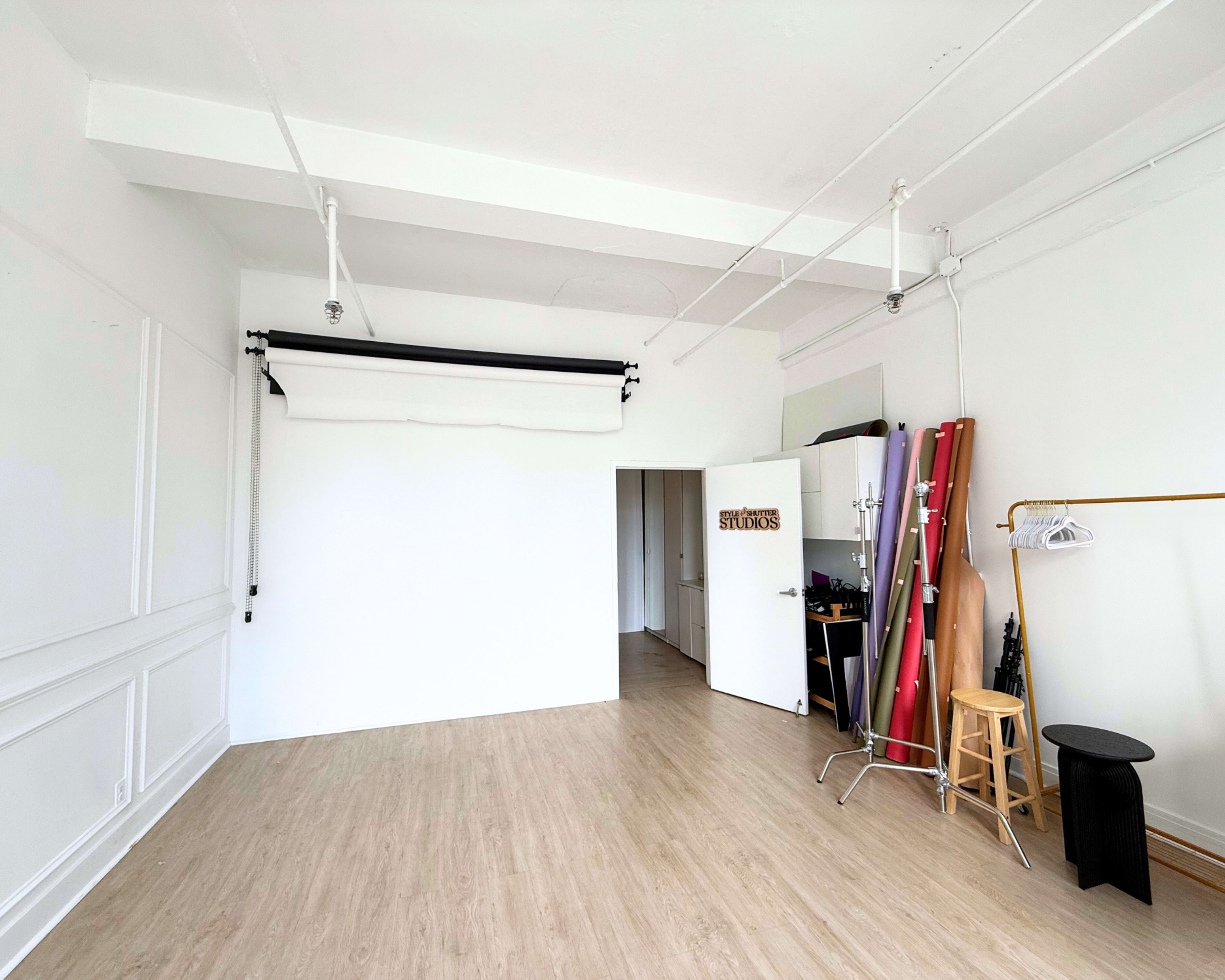 The image shows an empty studio space with a light wood floor, a backdrop setup on the left, a rack for rolls of paper in the center, and a door labeled "STUDIOS" on the back wall.
