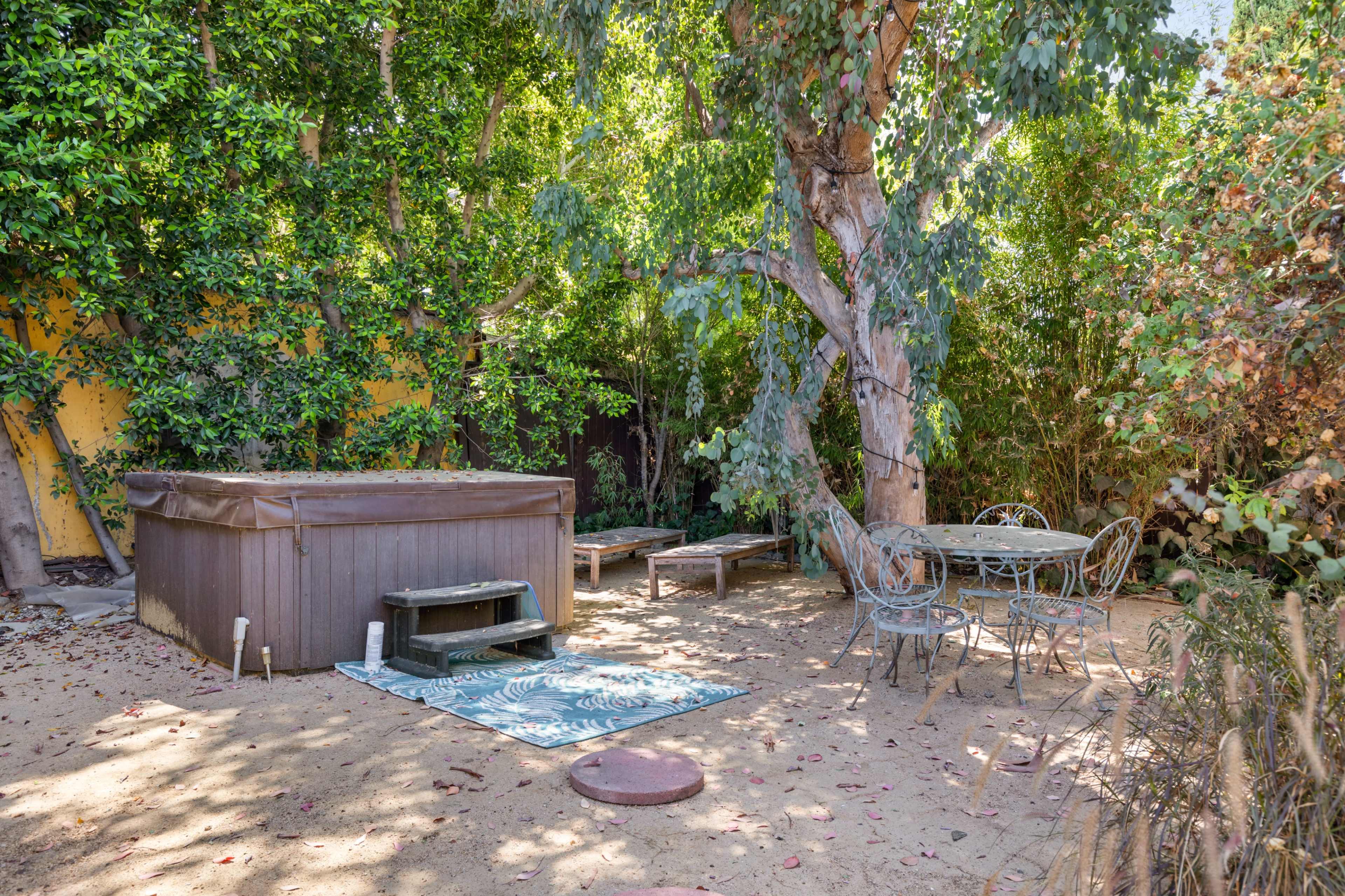 A secluded backyard area with a hot tub, a wooden bench, a circular dining table with metal chairs, and a large tree surrounded by greenery.