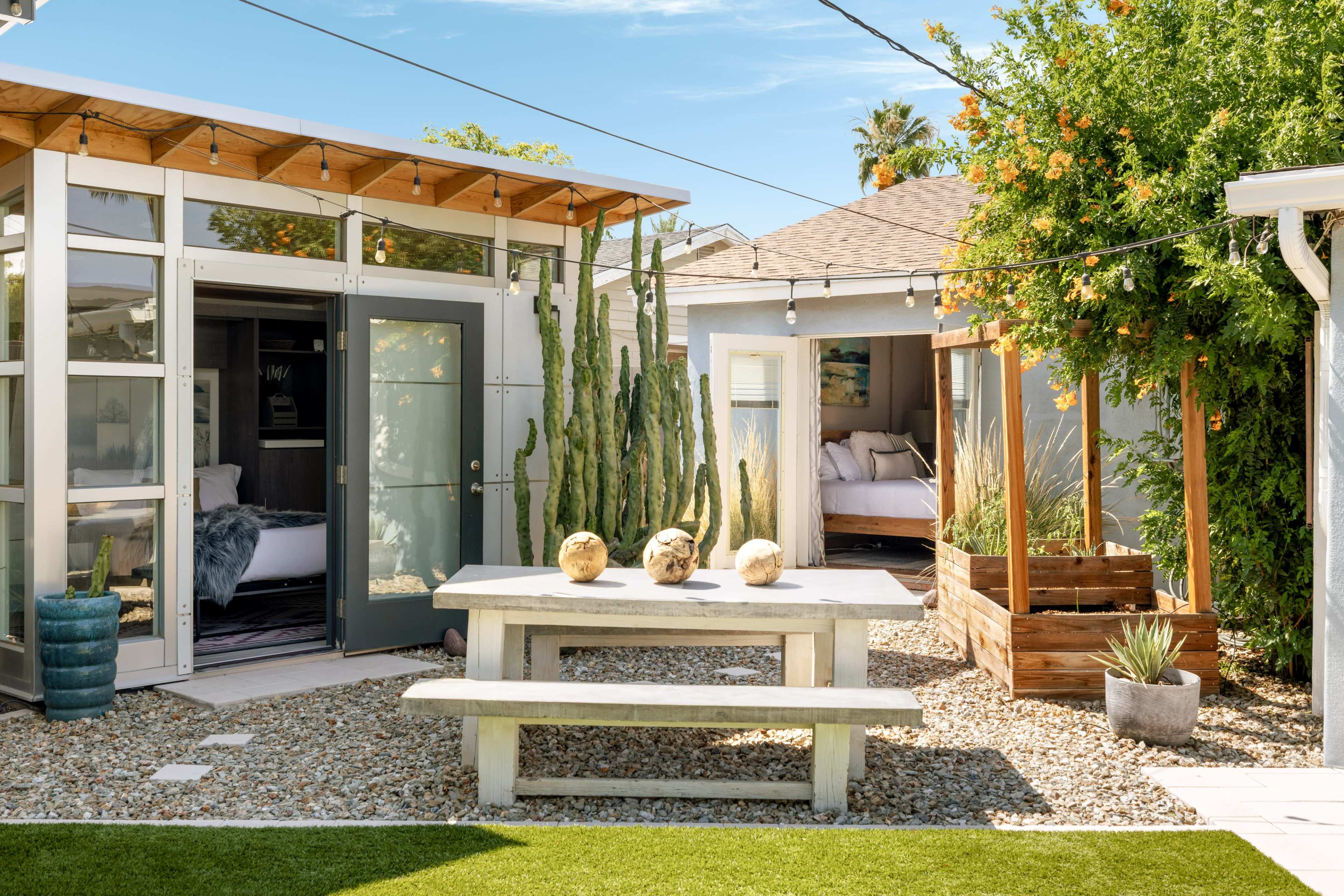 A modern outdoor courtyard features a wooden table with three decorative items, surrounded by cacti, gravel, and adjacent structures with large windows.