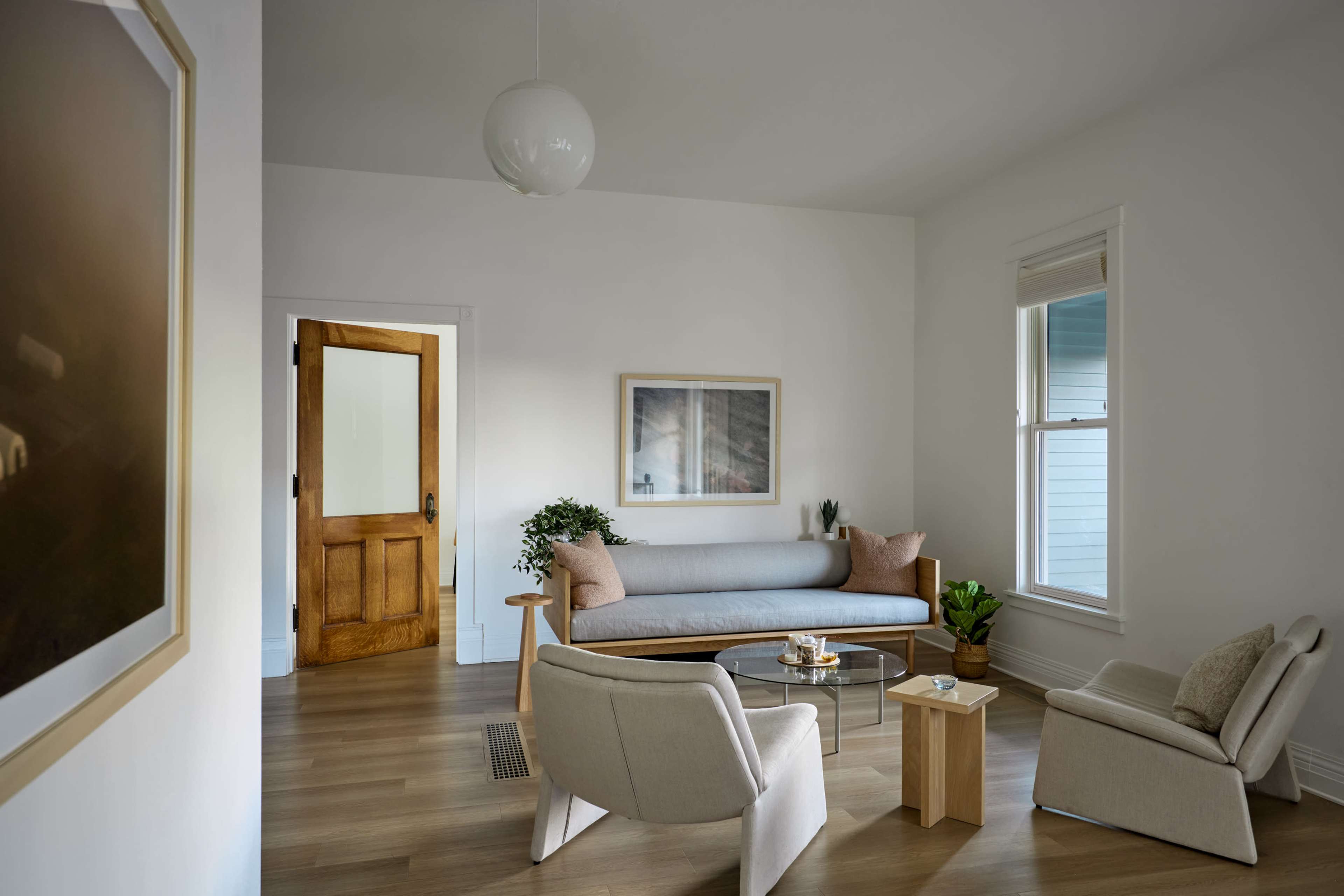 The image shows a modern living room featuring a gray sofa, two light-colored armchairs, a glass coffee table, and a wooden door leading to another room.
