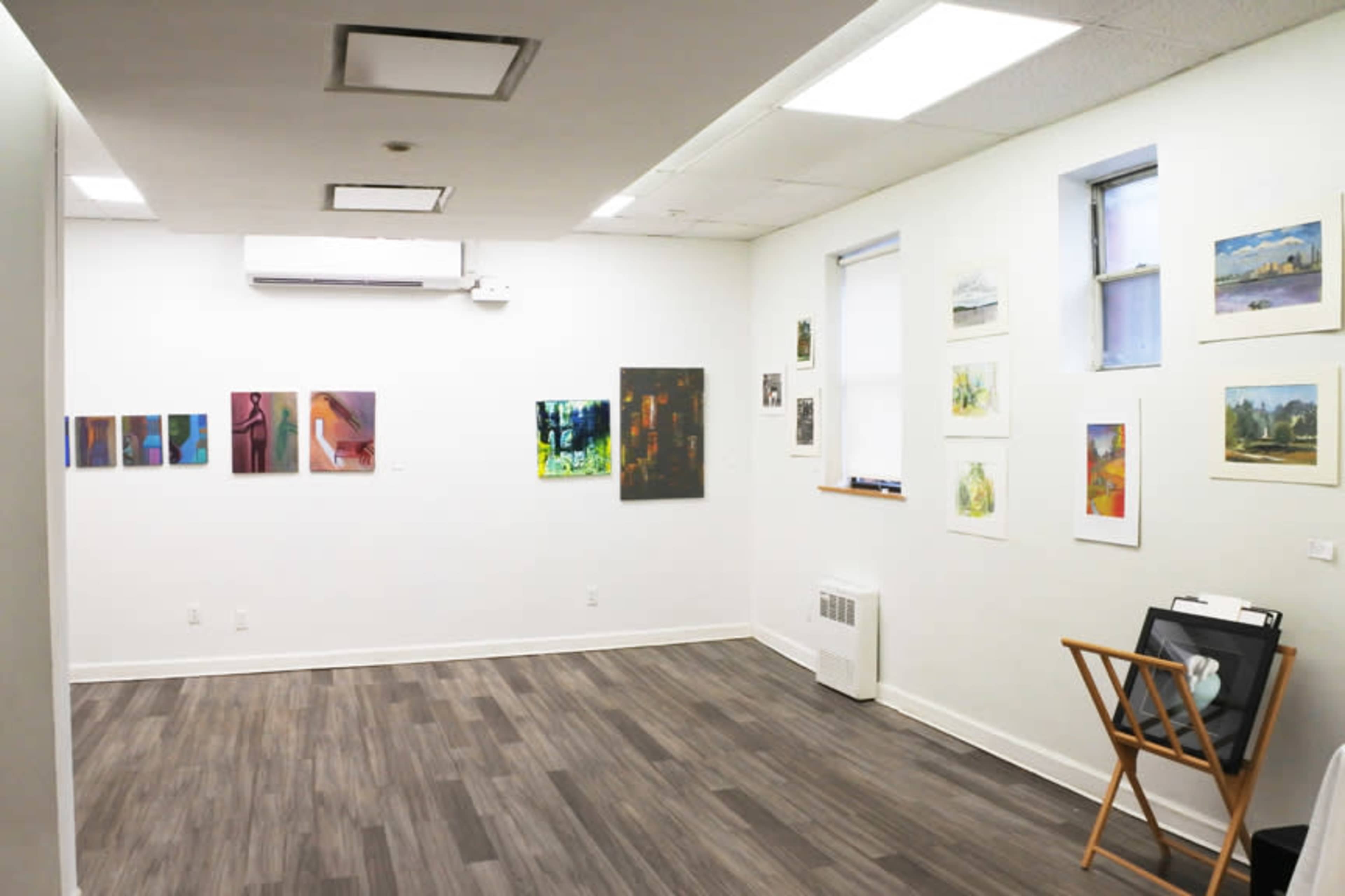 The image shows an art gallery with various paintings and artworks displayed on the walls and a folding chair in the corner.