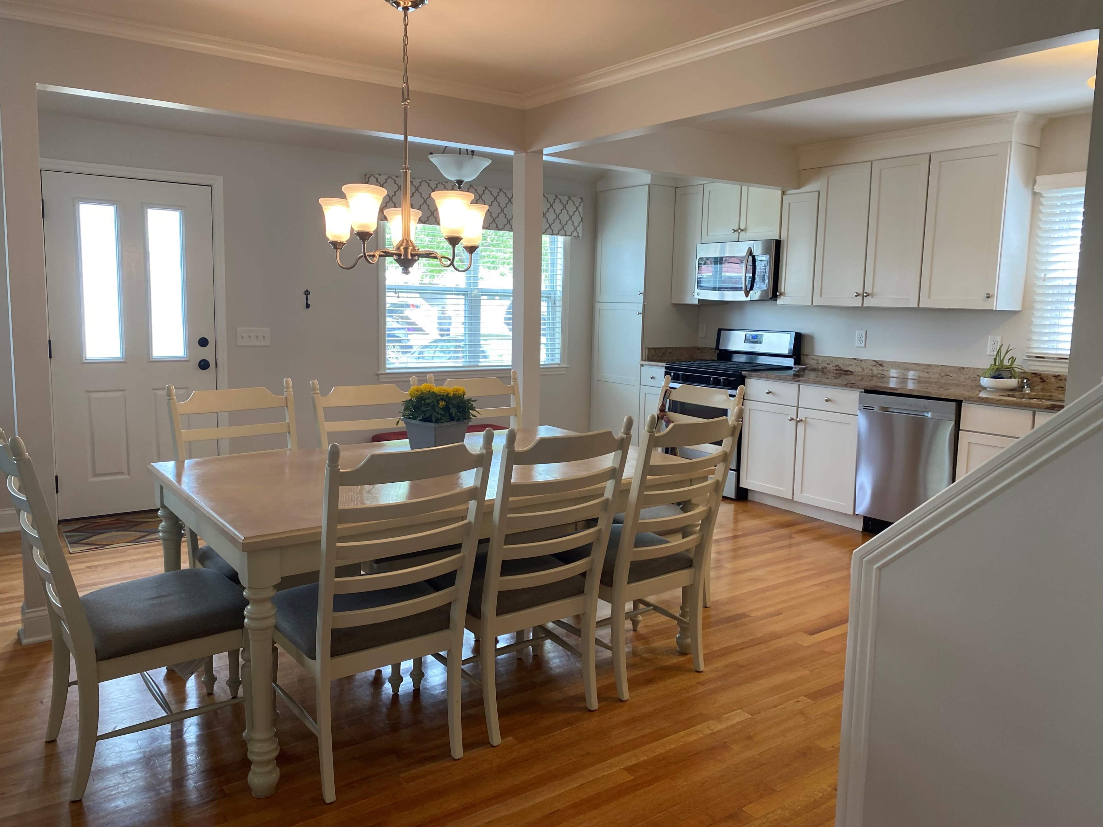 A well-lit dining area featuring a large white wooden table surrounded by chairs, with a view of a modern kitchen and an entrance door in the background.