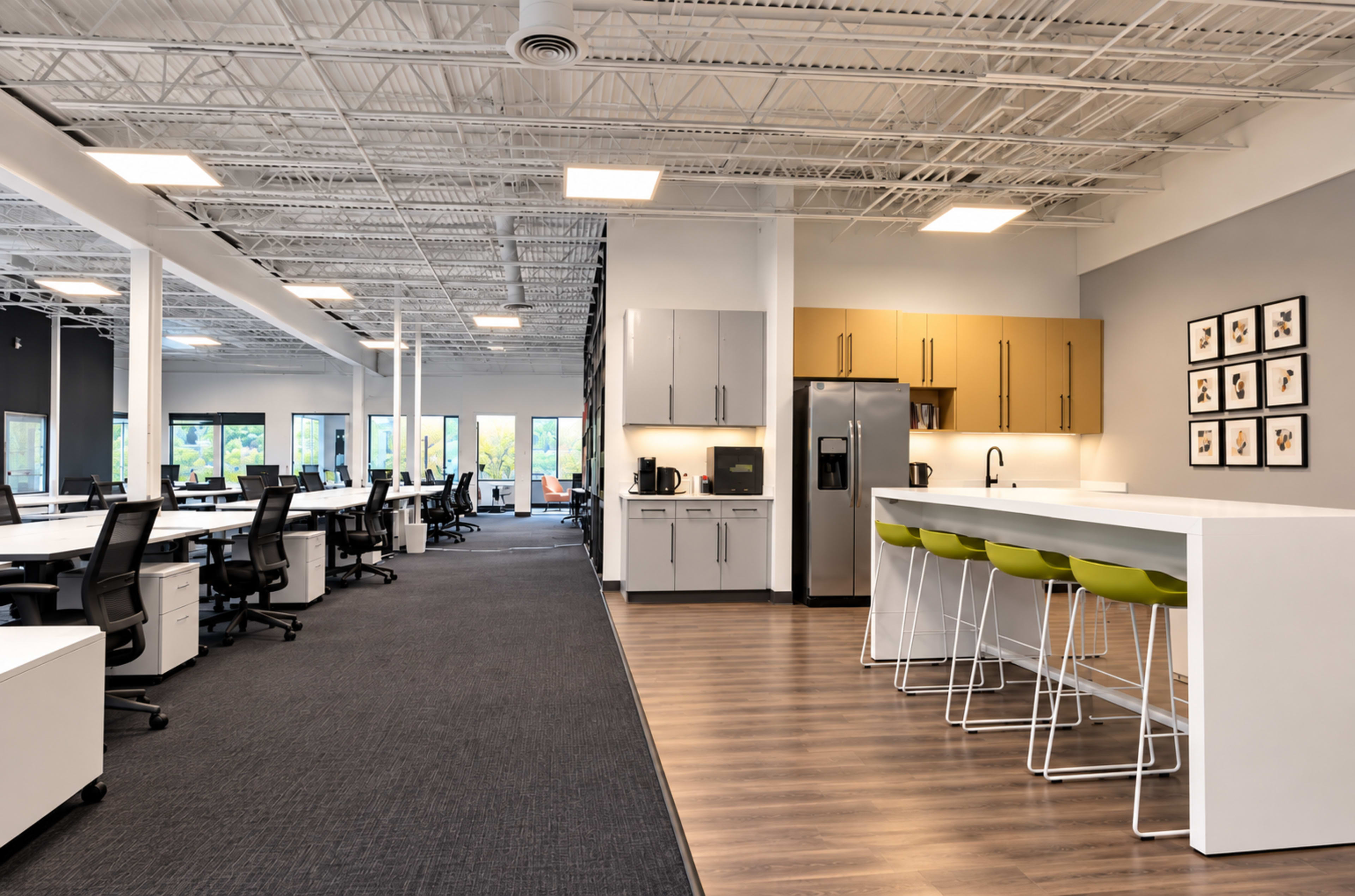 The image shows a modern office space featuring a kitchen area with bar stools and a work area with several desks and chairs.