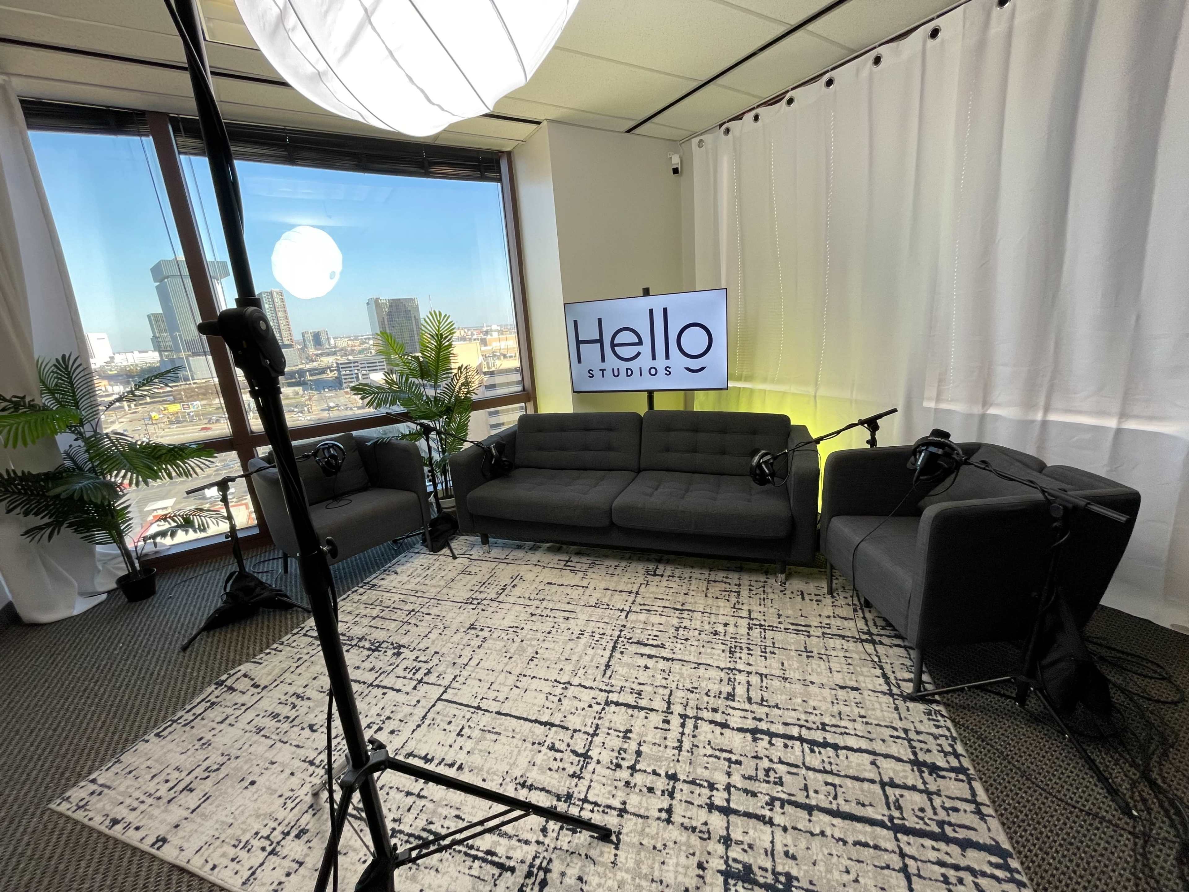 The image shows a neatly arranged studio setting with a grey couch and two armchairs, a large screen displaying "Hello Studios," and a window view of a cityscape.