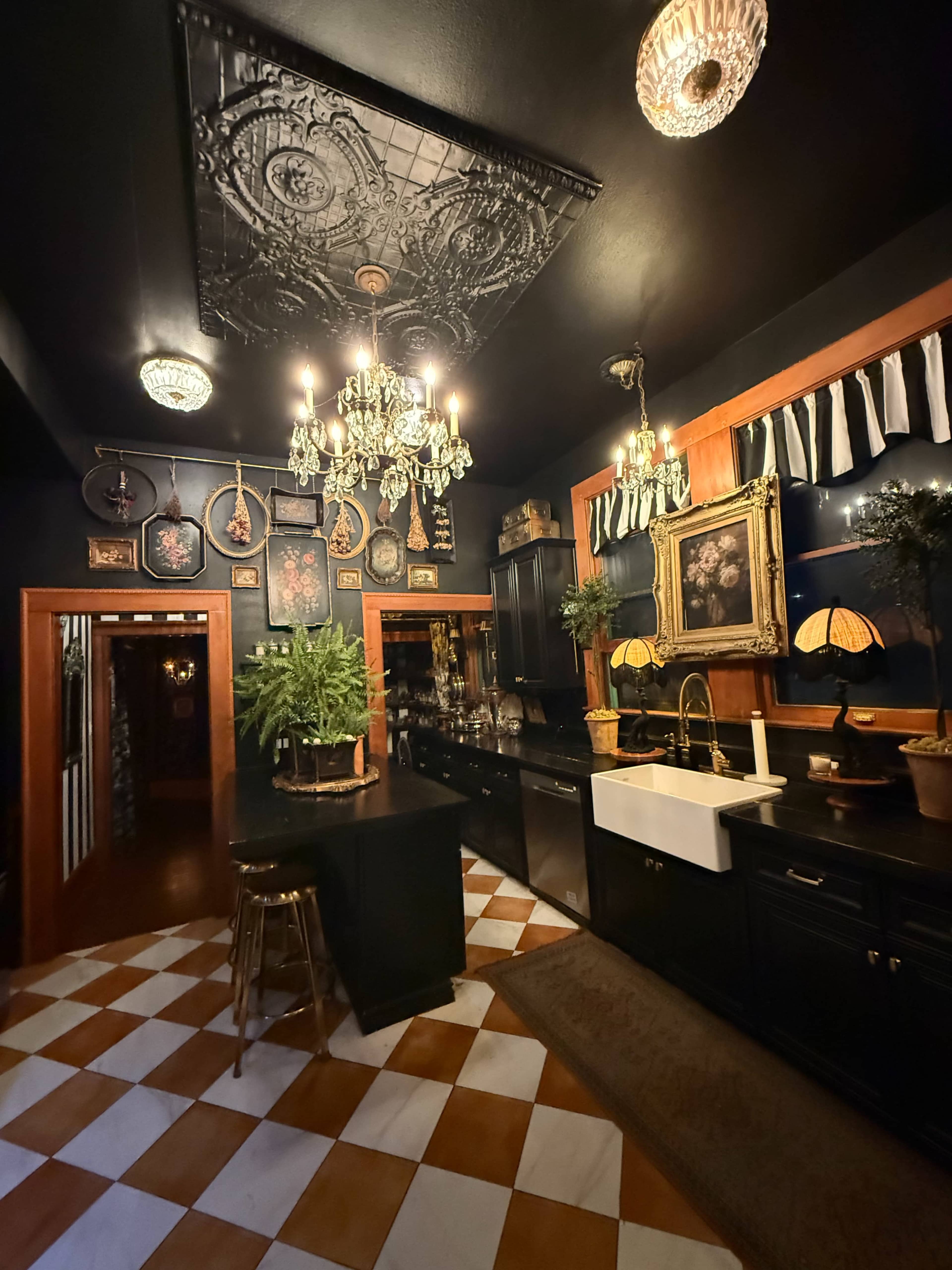 The image shows a stylish kitchen with black cabinetry, a farmhouse sink, and a patterned tile floor, featuring decorative elements like chandeliers and plants.